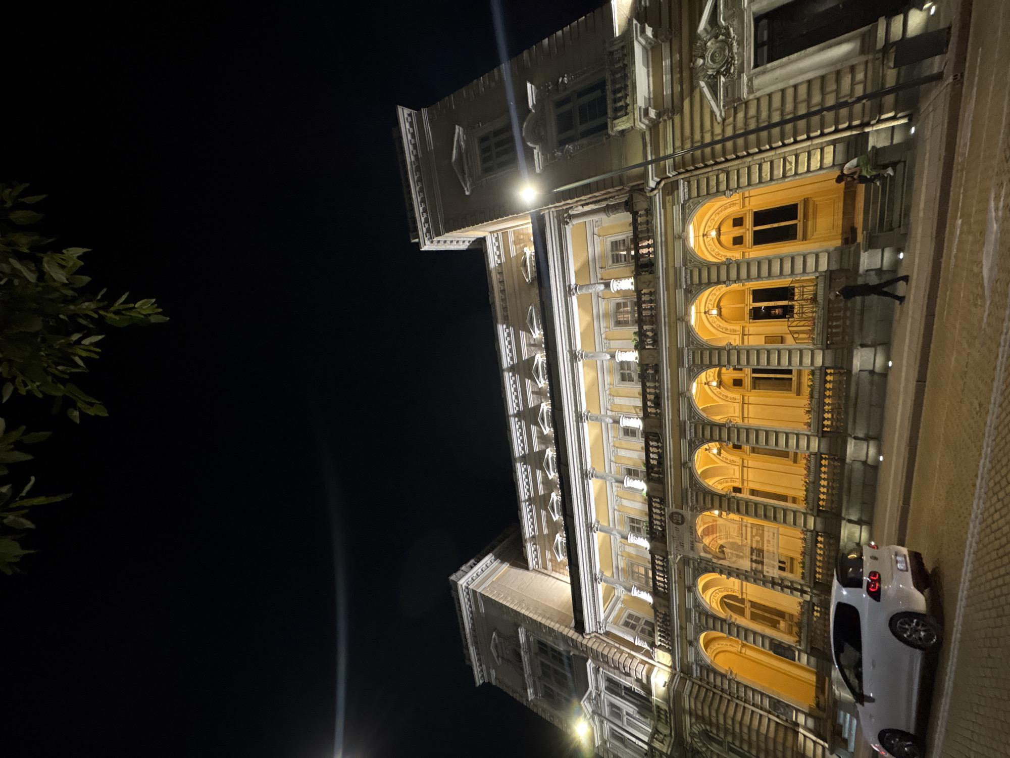 The image presents a vibrant nighttime scene of a historic, multi-story building, likely in Sofia, Bulgaria. The building's ornate facade is brightly illuminated with warm yellow and white artificial lights, highlighting its architectural details against a dark night sky.
The building features several large arched windows or entrances on its lower floors, glowing with a soft, warm light from within. Above these, multiple rectangular windows, some with closed shutters, are visible. The architecture includes decorative elements such as classical columns, balustrades, elaborate moldings, and cornices, suggesting a grand, possibly late 19th or early 20th-century European style.
A paved street or sidewalk runs alongside the building. Two individuals are present: one person in dark clothing walks along the pavement, while another, wearing a lighter top (possibly green or white), stands near one of the brightly lit arched doorways. A white hatchback car, resembling a Volkswagen Polo, is parked on the street next to the building.
In the upper left corner, green foliage from a tree or bush is visible against the dark sky. Within one of the arched window displays, a poster or sign is discernible, showing the text "21.09 / 20.00," likely indicating a date and time for an event. A partial Cyrillic word, "излож" (izlozh), hints at "изложба" (exhibition). The scene captures an active urban evening, with the beautifully lit architecture creating a striking contrast against the dark surroundings.