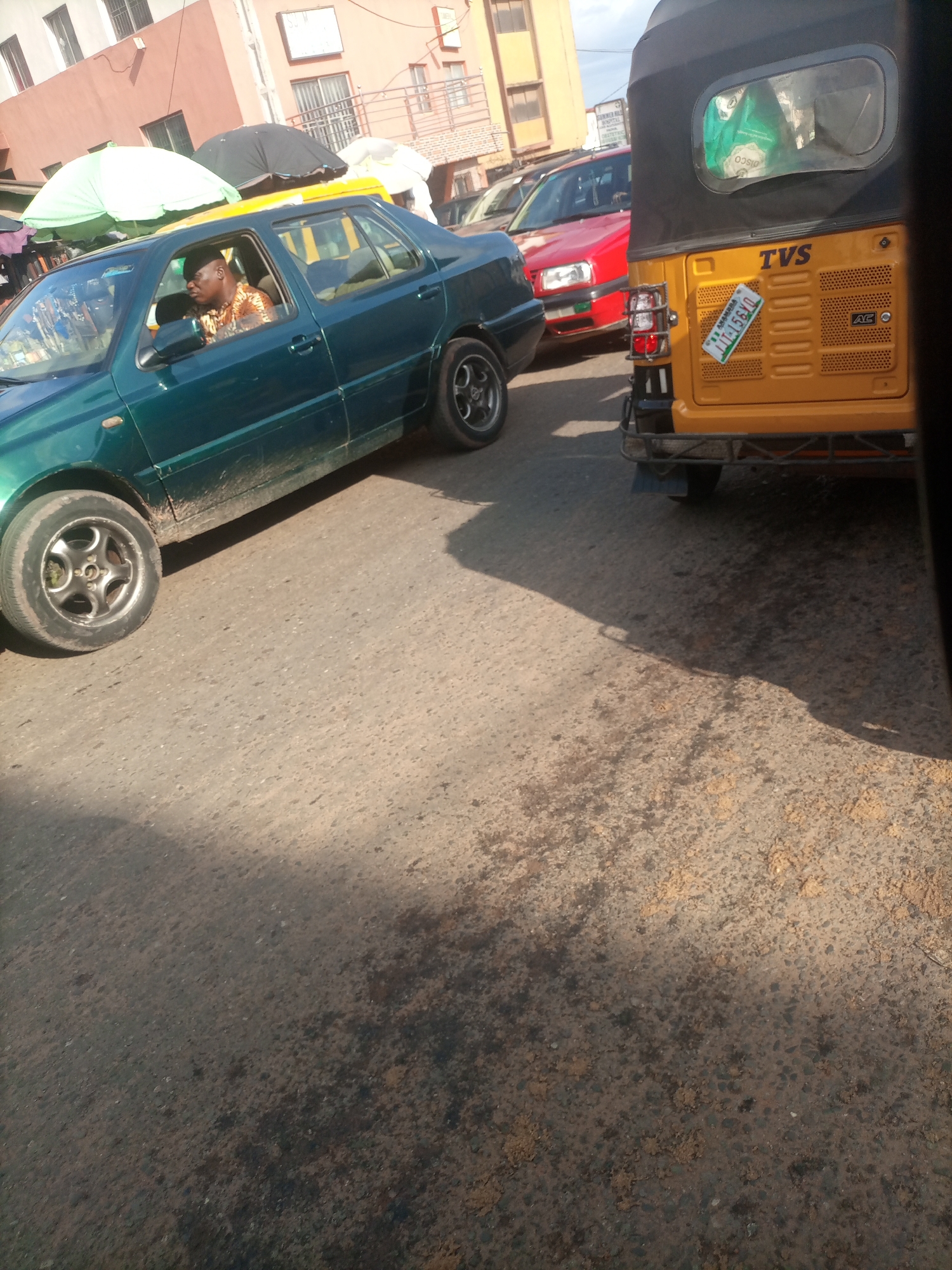 In Nkpor, Nigeria, a man in a patterned shirt drives a green sedan. The car is on a street, indicated by the asphalt road surface and the presence of other vehicles. To the right, a yellow auto-rickshaw, also known as a Keke Napep, is parked. The auto-rickshaw has a black roof and a license plate with the numbers "11-15610" and "ANAMBRA". In the background, there are buildings, suggesting an urban or semi-urban environment. The scene appears to be during the daytime, with shadows visible on the road.
