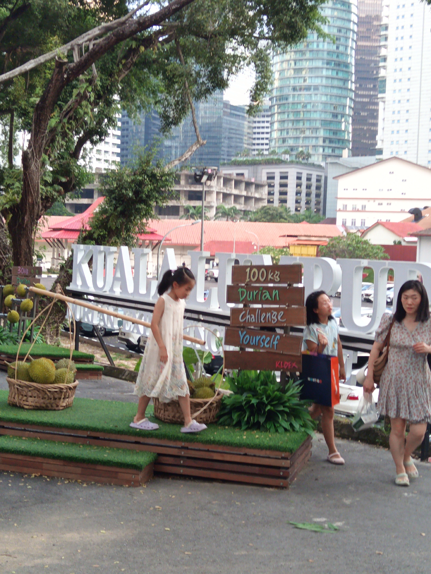 In Kuala Lumpur, Malaysia, a young girl in a white dress walks past a basket of durians. Behind her, a sign reads "100kg Durian Challenge Yourself." In the background, the iconic Petronas Twin Towers loom over the city skyline, with modern skyscrapers and traditional red-roofed buildings interspersed. The scene captures a vibrant street market or festival, with an emphasis on local produce and cultural attractions.