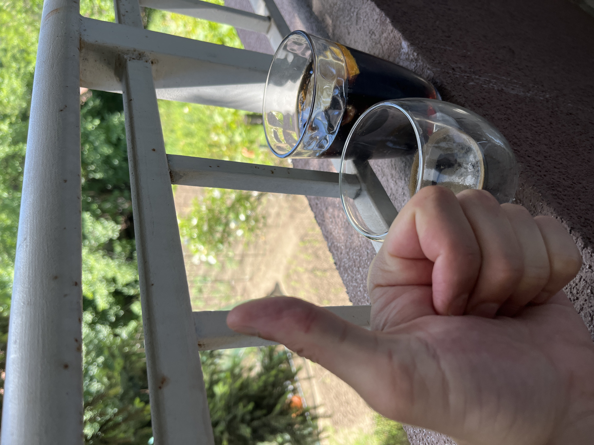 A close-up, eye-level shot shows a person's right thumb giving a thumbs-up gesture, positioned in the foreground and to the left. Behind the thumb, two glasses containing dark liquid, likely iced coffee or soda, rest on a concrete ledge. One glass is taller and appears to be a cola with ice and a slice of orange, while the other is shorter and wider, containing a lighter brown liquid with froth. The glasses and ledge are situated on a balcony or windowsill with a white railing in the mid-ground. Beyond the railing, a blurred background of green foliage and trees suggests an outdoor setting, possibly in a garden or park. The lighting indicates it is daytime, with sunlight casting shadows and highlighting the condensation on the glasses. The image composition focuses on the gesture and the drinks, with the background serving as a soft, natural backdrop. The overall scene evokes a sense of relaxation and enjoyment of a beverage outdoors. This image is not definitively set in Sofia, Bulgaria, and lacks specific landmarks or identifying features to confirm such a location.