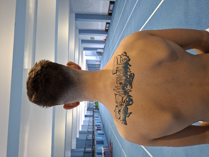 An adult male is depicted from behind, centered in the frame. He has short, dark brown hair with a faded cut on the sides and back. A large, stylized graffiti-style tattoo is centered on his upper back, spanning horizontally across his shoulder blades. The tattoo features the word "SOFIA" rendered in blue and black tones, accompanied by additional abstract graphic elements.
The subject stands on a blue indoor athletic track marked with white lane lines. The environment is a brightly lit indoor sports facility characterized by light blue walls, white structural beams, and a high ceiling. In the distant left background, track hurdles are visible. Along the right side of the track, further back, wooden benches are present. The specific location is identified as the Asics Arena in Sofia, Bulgaria.