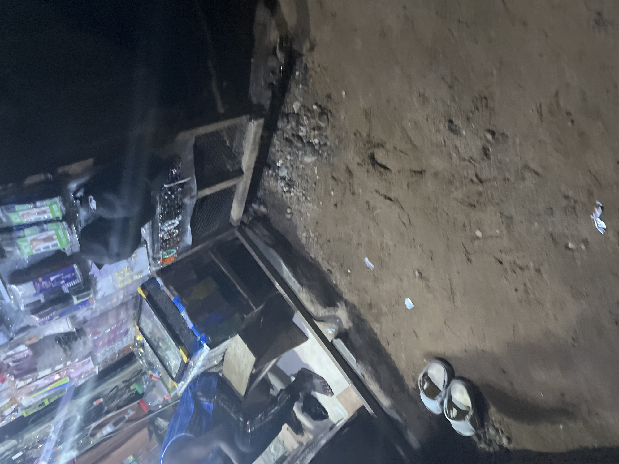 The image depicts an outdoor stall at night, possibly in a market. The stall is stocked with various items, including what appear to be phone accessories, jewelry, and other small goods displayed in plastic packaging. A person, partially visible, is seated at the stall wearing dark clothing and possibly black leggings. Their feet are bare, with one foot resting on the ground. In the foreground, on the dirt ground, a pair of white flip-flops are visible. The ground is uneven and appears to be a mix of dirt and small stones. The lighting suggests artificial light sources illuminating the stall. The overall scene is dark with limited visibility in some areas.