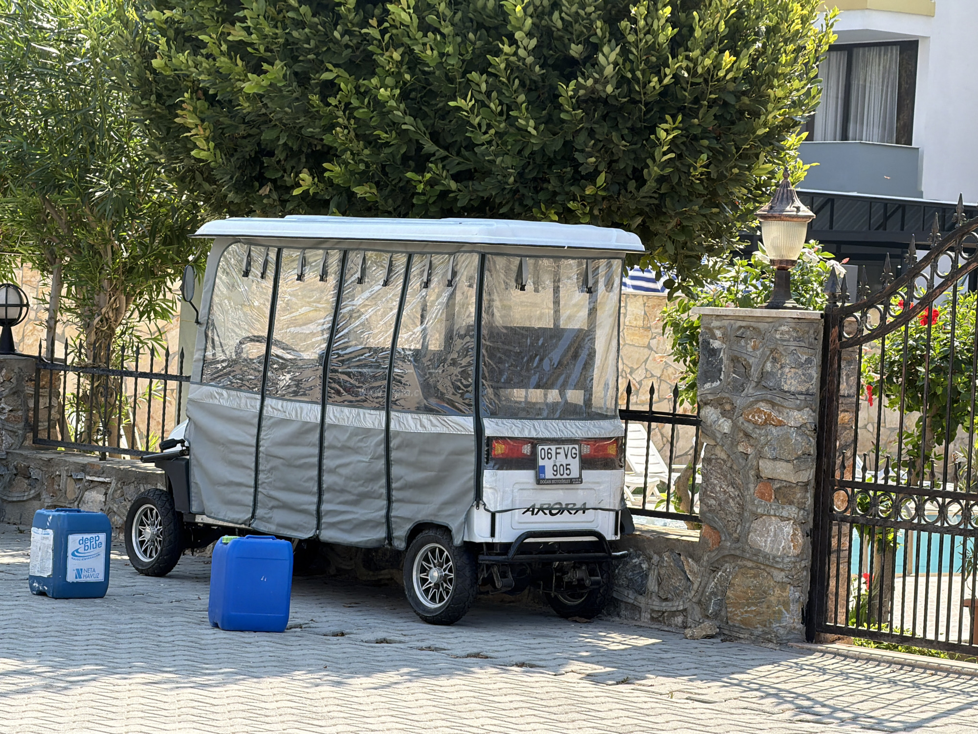 In Alanya, Turkey, a white golf cart with a grey tarp cover is parked on a paved surface. The golf cart has clear plastic sides, revealing a seating area inside. A license plate on the back reads "06 FVG 905". Two blue plastic containers are on the ground in front of the golf cart. To the right of the golf cart, a black wrought-iron gate with decorative circles stands open, revealing a swimming pool with blue water. A stone wall is visible behind the gate and extends along the side of the property. Lush green trees and foliage surround the scene, with a modern building visible in the background. A vintage-style lamp post is mounted on the stone wall near the gate.