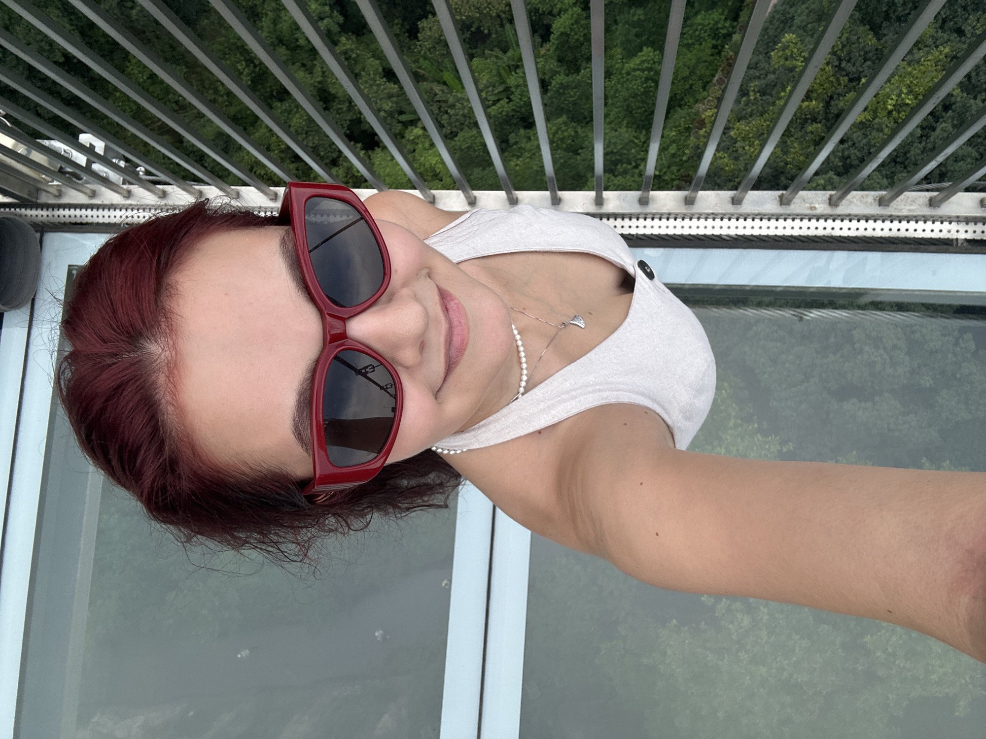 A young woman with red hair and red-rimmed sunglasses is taking a selfie from a high vantage point, possibly a glass-floored observation deck. She is wearing a white tank top and a pearl necklace with a pendant. To her right, a metal railing separates her from a dense, green forest below. The woman is smiling and looking at the camera. The image appears to be taken at the Qingyuan Sky Tower in Qingyuan City, China, known for its glass-bottomed observation deck offering panoramic views of the surrounding landscape.