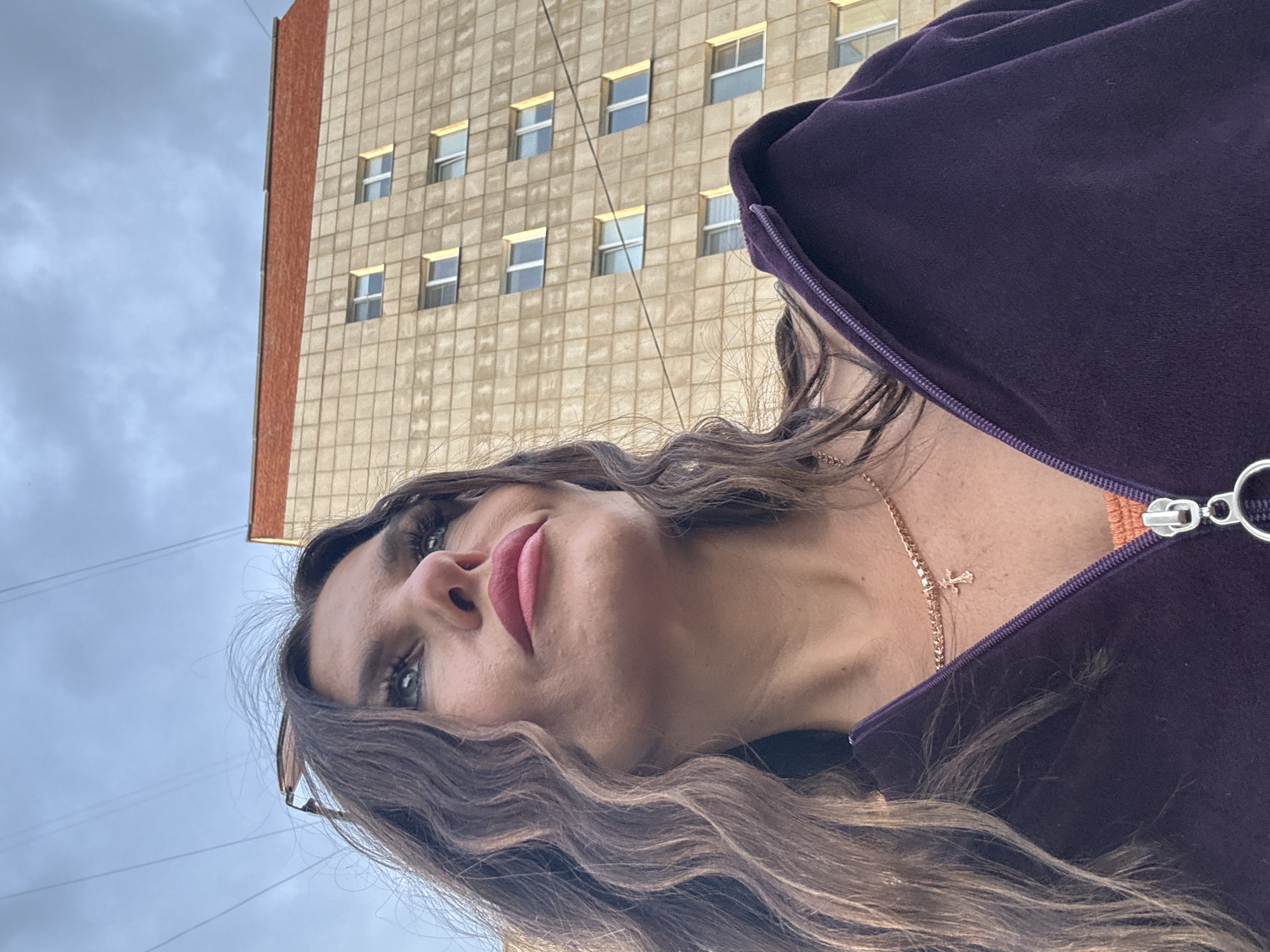 This media file features a close-up, low-angle shot of a woman in Chaikovsky, Russia. The woman has long, wavy brown hair and is wearing a purple zippered jacket over an orange garment. She is also wearing a delicate gold necklace with a small cross pendant. Her makeup includes dark eyeliner and pink lipstick, and she is looking upwards and to her right.
In the background, a light-colored, multi-story building with a reddish-brown accent strip is visible. The building has numerous windows, many of which are illuminated. The sky is overcast and cloudy, suggesting a daytime setting, possibly late afternoon or early evening given the lighting on the building. The image is taken outdoors, with power lines visible against the sky. The overall mood appears casual and candid. There is no visible text in the image.