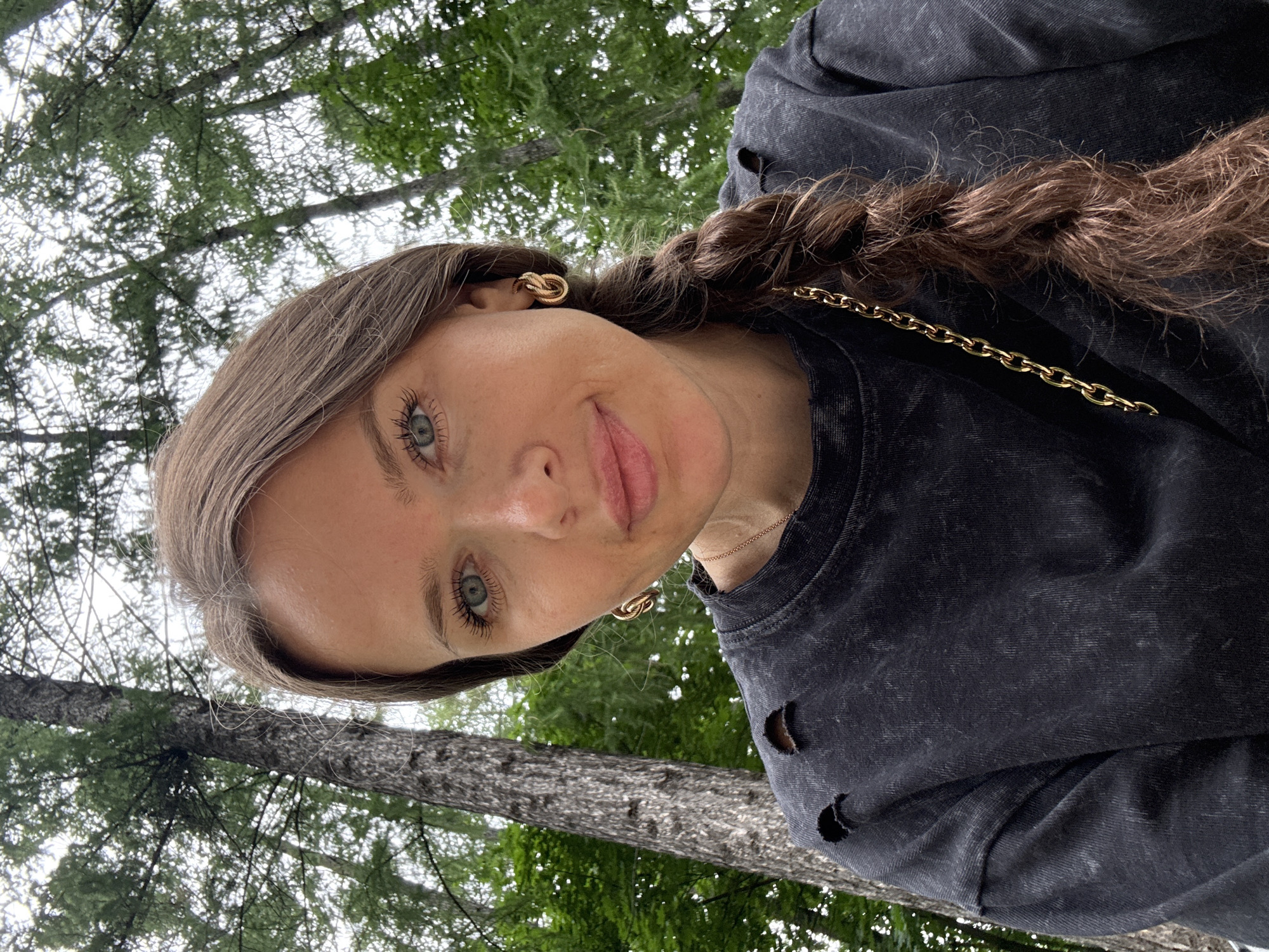 The image is a close-up, rotated selfie of a young woman outdoors, with trees and foliage in the background. She has long, brown hair braided and draped over her shoulder. She is wearing a dark, distressed sweatshirt and a gold chain necklace. Her eyes are a striking blue-green, and she is looking towards the camera with a slight smile. A gold, knot-shaped earring is visible in her left ear. The scene appears to be set in a natural environment, possibly a park or forest, with dappled light suggesting it is daytime. The overall mood is casual and relaxed. No visible text is present.