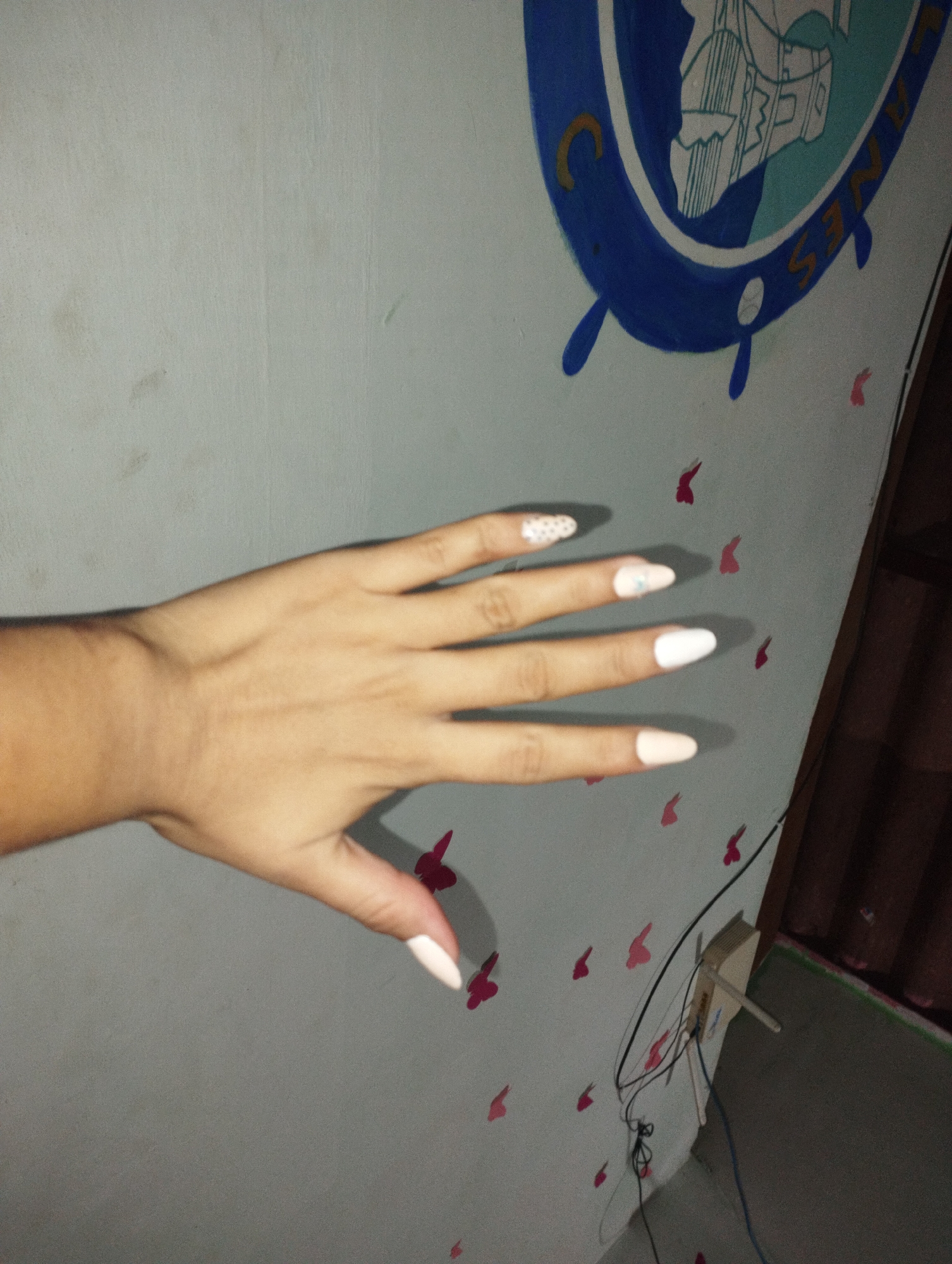 A person's left hand with artificial fingernails is held up against a light gray wall. The fingernails are various shades of pink and white, with one nail adorned with small white dots and another with a subtle pink floral design. The wall has a painted blue and white emblem with gold lettering and several small red butterfly stickers. The scene appears to be indoors, possibly in Punta de Piedras, Venezuela, based on contextual information not visible in the image itself. The lighting suggests it is nighttime or a dimly lit room.