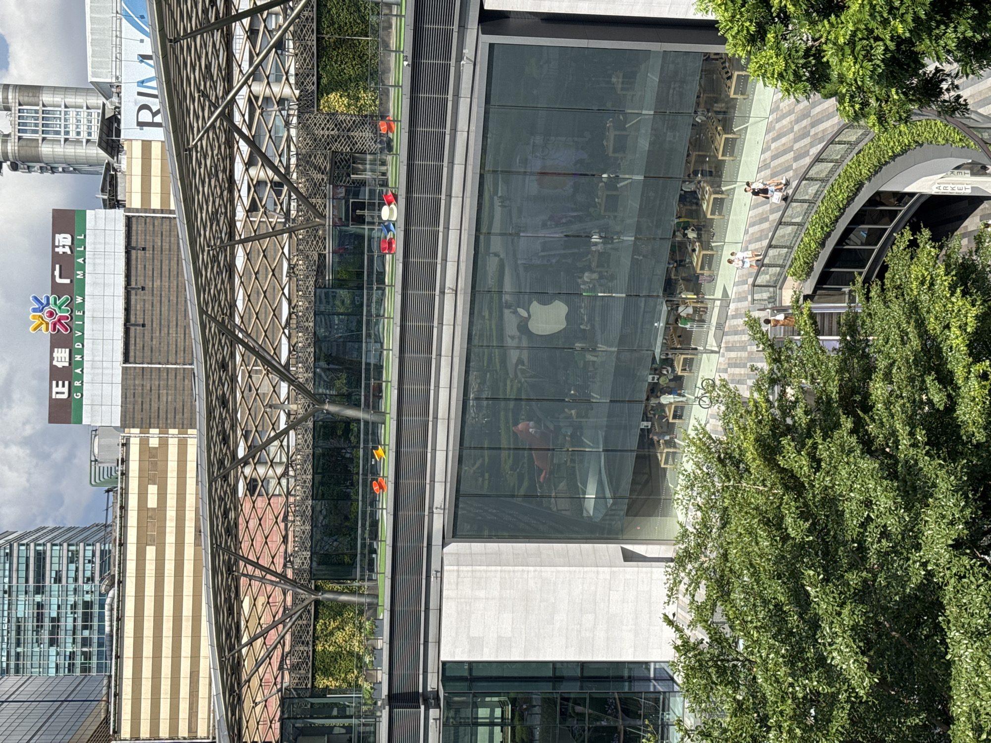 The image depicts an exterior view of a modern architectural space in the Tianhe District of Guangzhou, China. The focal point is a large glass-walled Apple store, recognizable by its prominent white Apple logo. The store's interior is visible through the glass, revealing tables, chairs, and a number of people inside. The store's architecture includes an integrated outdoor area with landscaping, including a walkway paved with a geometric pattern. Several people are observed walking or standing in this area. The surrounding environment is densely urban, with several tall buildings visible in the background. One building is identified by the name "Grandview Mall." A complex metal structure, possibly a canopy or architectural element, extends over part of the scene. Trees are also present, partially obscuring the view and adding greenery to the urban landscape.