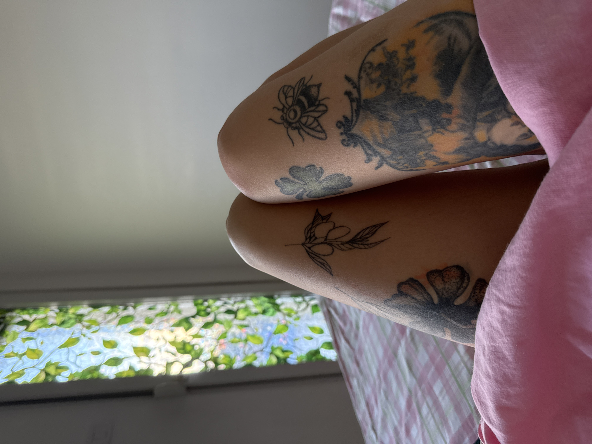 The image shows a close-up view of a person's legs, which are covered in various tattoos. The tattoos include a branch with olives, a bee, a clover, and a larger, more complex design that appears to be a portrait or scene. The legs are resting on a pink blanket, and the background shows a bed with a pink and white checkered sheet. To the left, there is a window with a stained-glass effect, depicting green leaves and a blue sky. The lighting suggests it is daytime, possibly morning or afternoon.