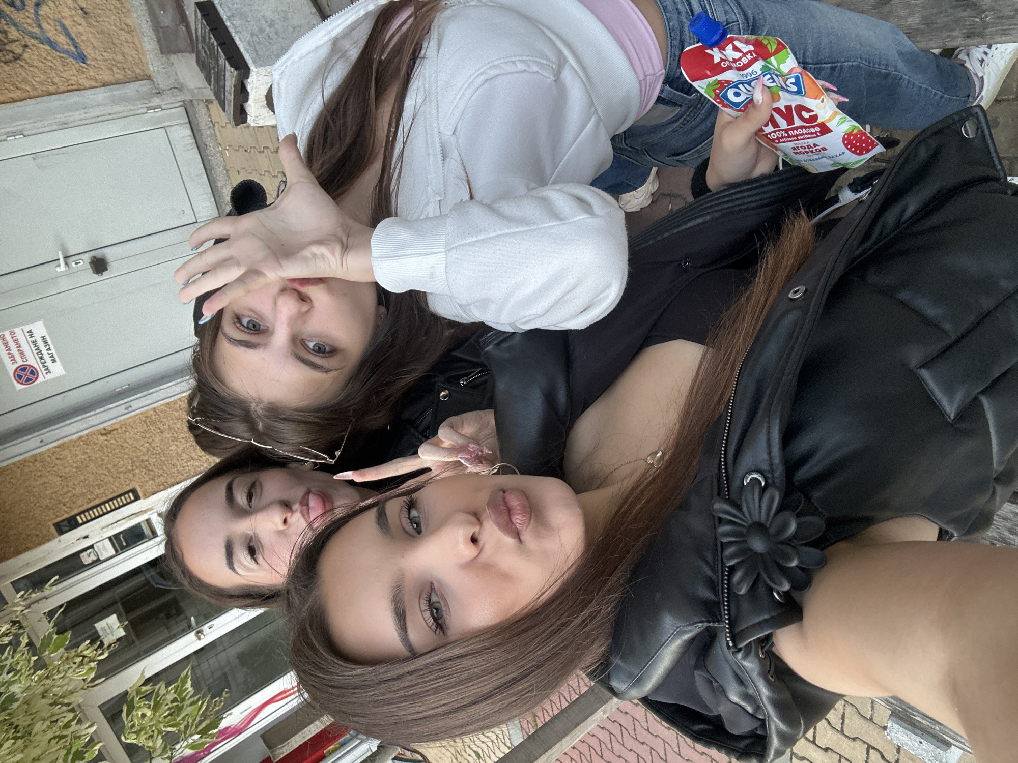 Three young women are taking a selfie outdoors in front of a building. The woman in the foreground on the left has long brown hair, is wearing a black leather jacket, and is puckering her lips. The woman in the middle has long brown hair and is sticking out her tongue. The woman on the right has long brown hair and is holding up her hand with her fingers spread. She is wearing a white hoodie and jeans. She is also holding a pouch of yogurt. The background shows a building with a gray door and a light brown textured wall. There are some trees and plants to the left of the building. The ground is paved with light brown bricks. The scene is located in the city of Shumen, Bulgaria.