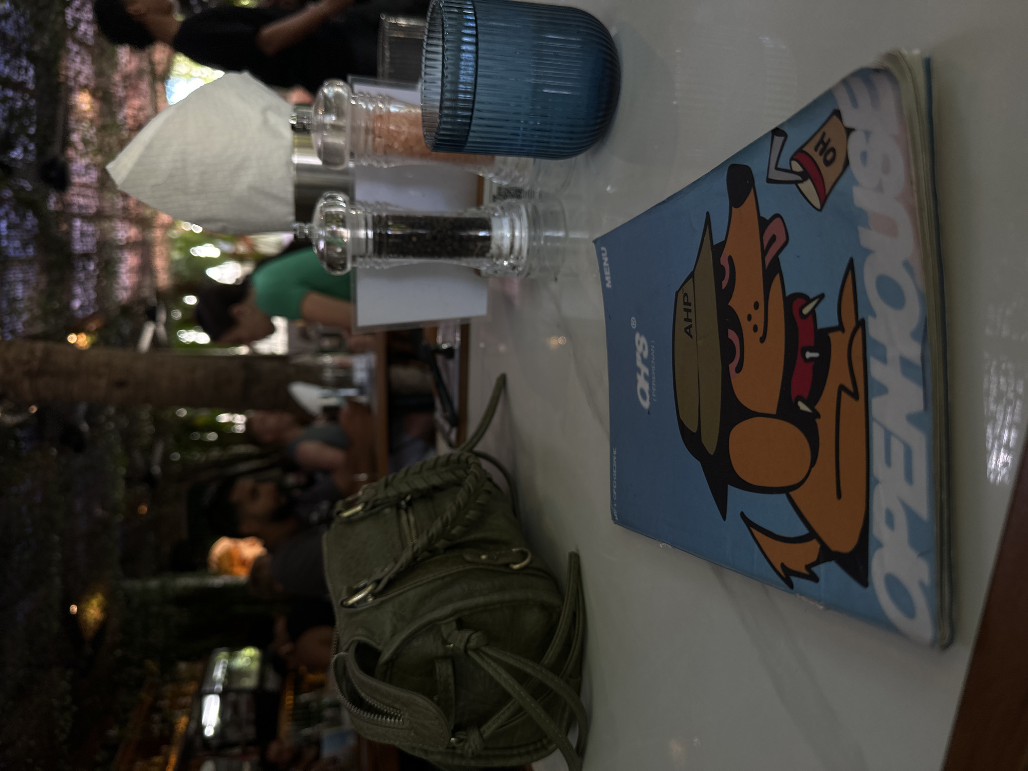 A table-top view shows a blue menu with a cartoon dog wearing a hat and sunglasses, holding a cup with "OH" on it. The dog's tongue is out, and it has a red collar. Below the dog, the word "OPENHOUSE" is prominently displayed. In the background, a green handbag with braided handles rests on the table. Two salt and pepper grinders are visible, along with a ribbed blue glass. The scene appears to be set in a restaurant with blurred figures of people dining and a bar area in the distance. The location is identified as OH's Bar & Grill, located in Mengwi, Indonesia.