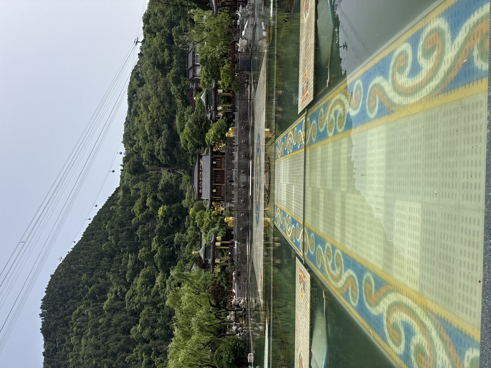 The image shows a landscape in Lintong District, China. A green mountain covered in trees is visible, with a cable car system running up its slope. At the foot of the mountain, there is a complex of traditional Chinese buildings, possibly a temple or historical site, surrounded by trees. A reflective surface, perhaps water or polished stone, creates a mirrored effect, doubling the appearance of the buildings and the mountain. A decorated walkway or platform extends from the buildings towards the viewer, featuring a blue and yellow swirling pattern. The sky appears overcast, contributing to a subdued and slightly muted color palette in the scene.