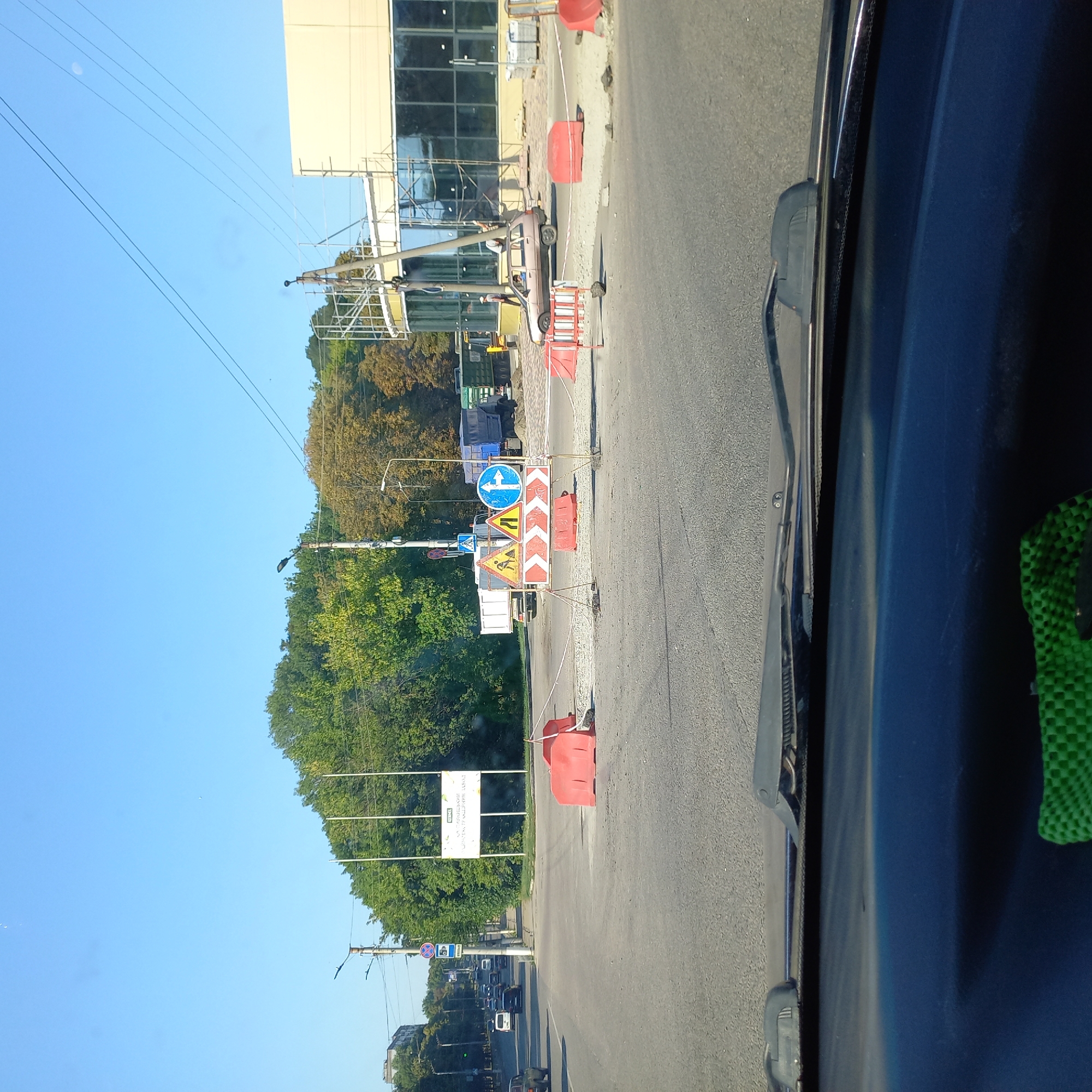 The image depicts a road construction site in Kropyvnytskyi, Ukraine. The scene is viewed from inside a vehicle, looking forward through the windshield. In the foreground, the road surface shows signs of ongoing work, with red traffic barriers and white tape delineating the construction area. Several construction signs are visible, including a warning sign for road work, a sign indicating a narrow road, and a blue directional sign with a white arrow pointing right and a white line indicating a right turn.
Further ahead, traffic signs indicate a roundabout or intersection. Trucks, including a white dump truck and a blue truck, are parked or moving in the background. A light-colored car is also present, with a driver inside. On the right side of the image, a building under construction is visible, with scaffolding and a light-colored facade. Trees line the background, suggesting a somewhat urban or suburban environment. The sky is clear and blue, indicating daytime.
The specific location appears to be an intersection or a section of road undergoing significant repair or reconstruction, likely in a developed area of Kropyvnytskyi given the presence of buildings and established infrastructure. The overall impression is one of active construction work impacting traffic flow.