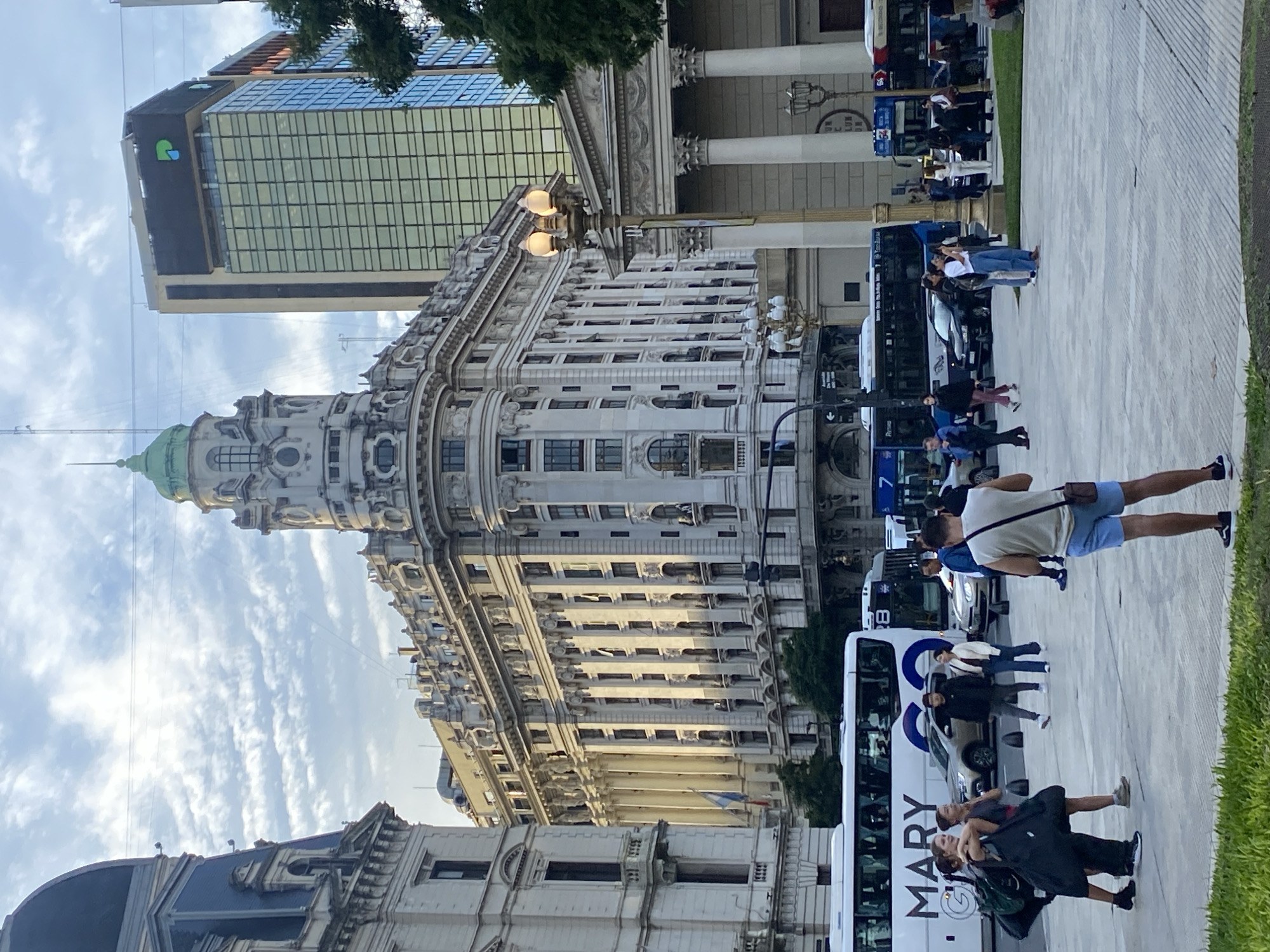 The image shows a cityscape in Buenos Aires, Argentina. The architecture is ornate, with a tall building featuring a green dome and elaborate decorations. Adjacent to it is a modern glass skyscraper with a green and blue logo. People are walking along a paved area beside the buildings. Buses are parked nearby, some with "Mary" displayed on their sides. The sky is partly cloudy.