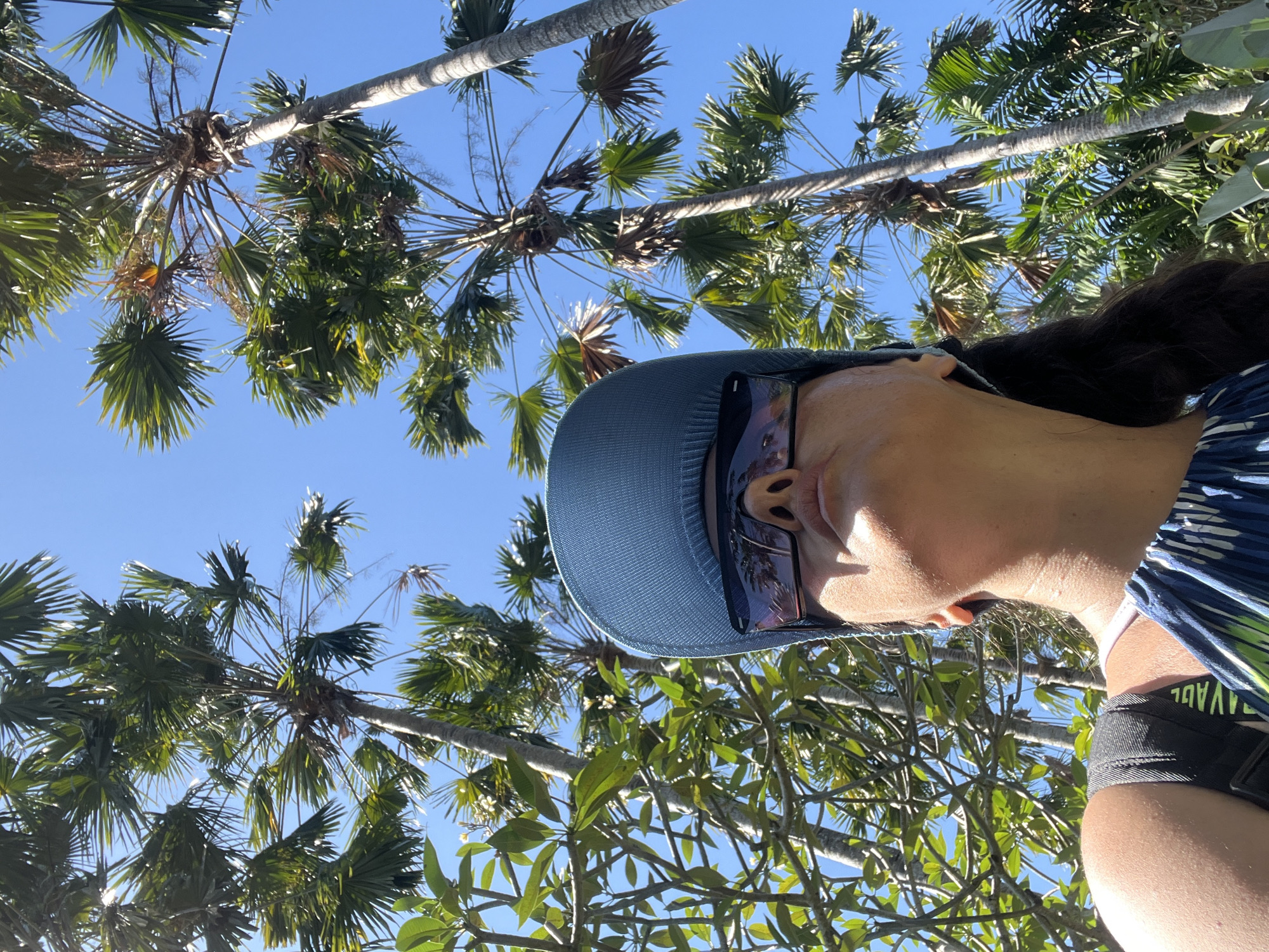 A low-angle, medium shot captures a person wearing a blue baseball cap and sunglasses, looking upwards. The individual's face is partially obscured by the accessories. They are situated in an outdoor environment characterized by lush greenery and tall palm trees under a clear blue sky. The person is wearing a blue and white striped top, and a black backpack strap is visible over their left shoulder. The scene appears to be in a tropical location, potentially the city of Kuta Selatan, Indonesia, given the presence of palm trees and the bright, sunny atmosphere. The lighting suggests it is daytime, with sunlight illuminating the scene.