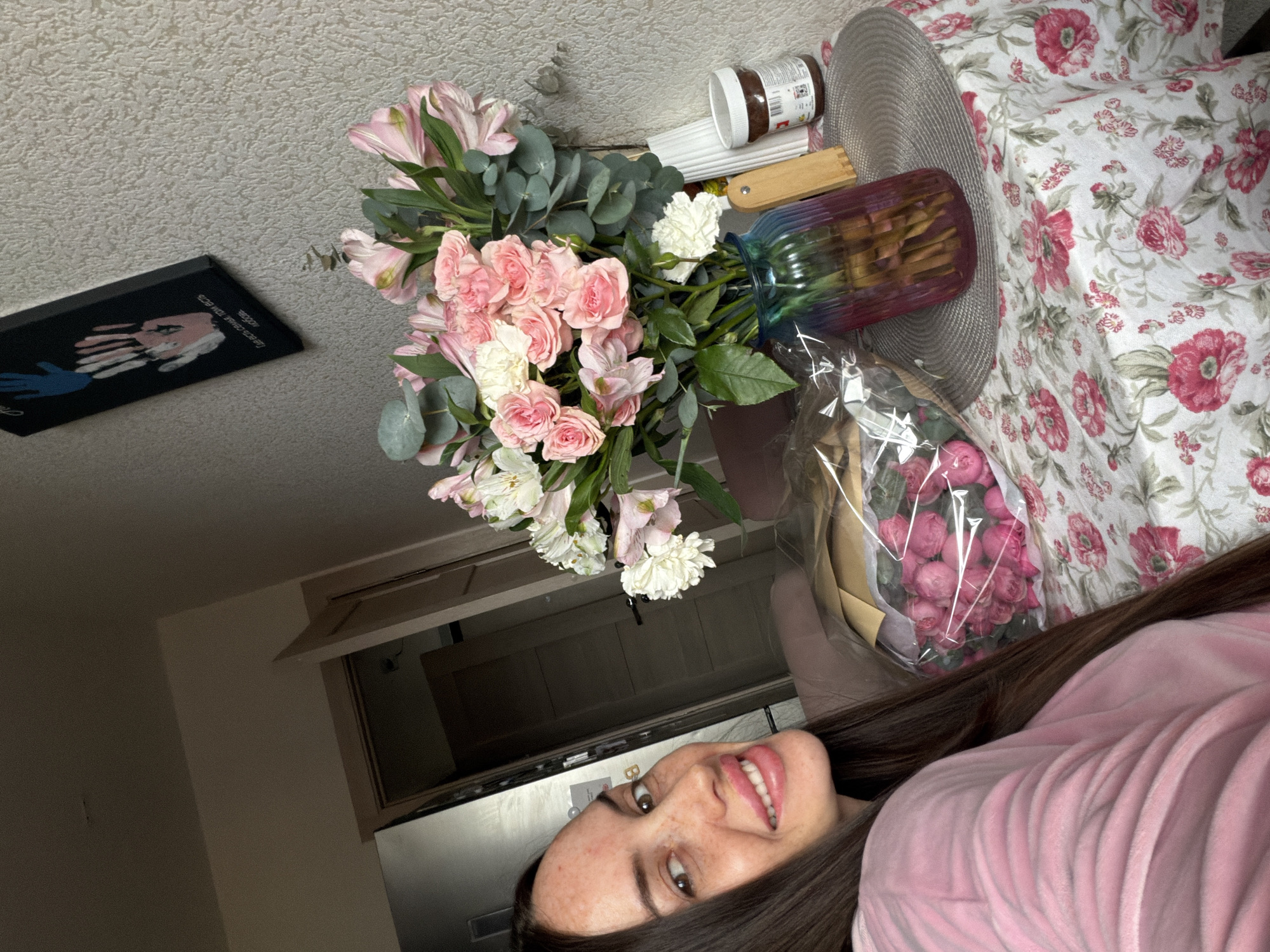 This is a media file featuring a young woman lying down, with her face prominently displayed in the foreground. She has dark hair, freckles, and is wearing a pink plush robe. She is smiling and appears to be in a relaxed or happy mood.
Surrounding her, and extending out of focus, are several objects. To her left, there is a bouquet of pink and white flowers, with eucalyptus leaves. Next to the flowers is a vase with a rainbow-colored glass design, containing what appear to be crackers or cookies. A jar of what looks like Nutella is also visible. In the background, there is a patterned fabric, possibly a blanket or tablecloth, with a floral design in pink and white. A dark framed picture with handprints is also visible on a textured wall. Further back, a cabinet or closet door is partially open, revealing darkness within.
The lighting suggests it is daytime, but the specific time of day is unclear. There are no obvious cues for weather. The setting appears to be indoors, likely a home environment. The overall impression is one of a casual, perhaps celebratory moment, given the presence of flowers and treats. There is no visible text that can be deciphered from the provided images.