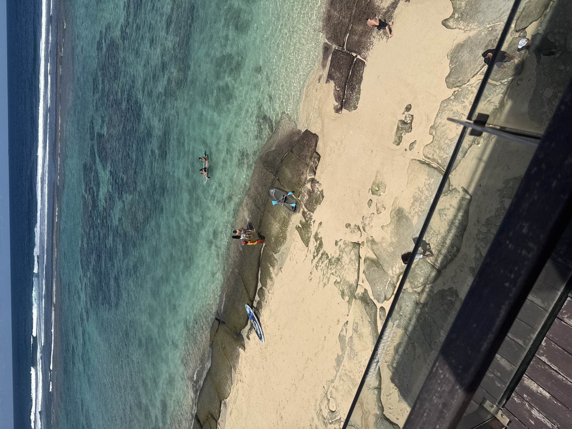 This photograph depicts an aerial view of a beach in Kuta Selatan, Indonesia. The water transitions from deep blue to turquoise as it approaches the shore, with visible waves breaking. The sandy beach is interspersed with rocky formations. Several people are visible: two individuals are in a clear kayak, and another group is standing on the beach near a blue and white paddleboard. A partially transparent kayak and paddles are situated on the beach. Another person is on the rocky formations further up the beach. The scene is viewed from an elevated position, possibly a pier or deck, with the railing partially visible in the foreground. The overall lighting suggests a sunny day.
