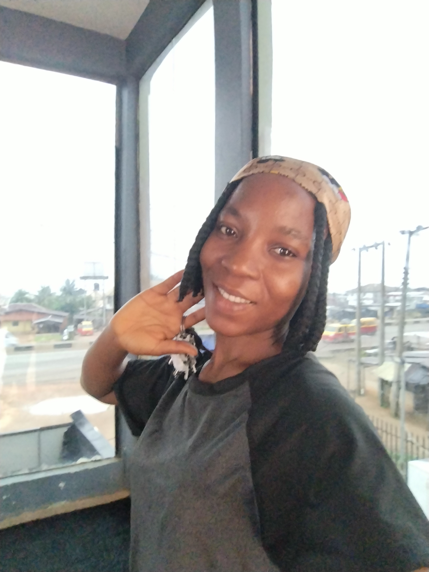 A young Black woman with braided hair and a headscarf smiles at the camera while standing in front of a large window. Her left hand is raised to her cheek, and she is wearing a dark t-shirt with grey raglan sleeves. The window offers a view of a street scene in Uselu, Nigeria, with buildings, trees, and vehicles visible. The overall lighting suggests it is daytime, likely overcast.
