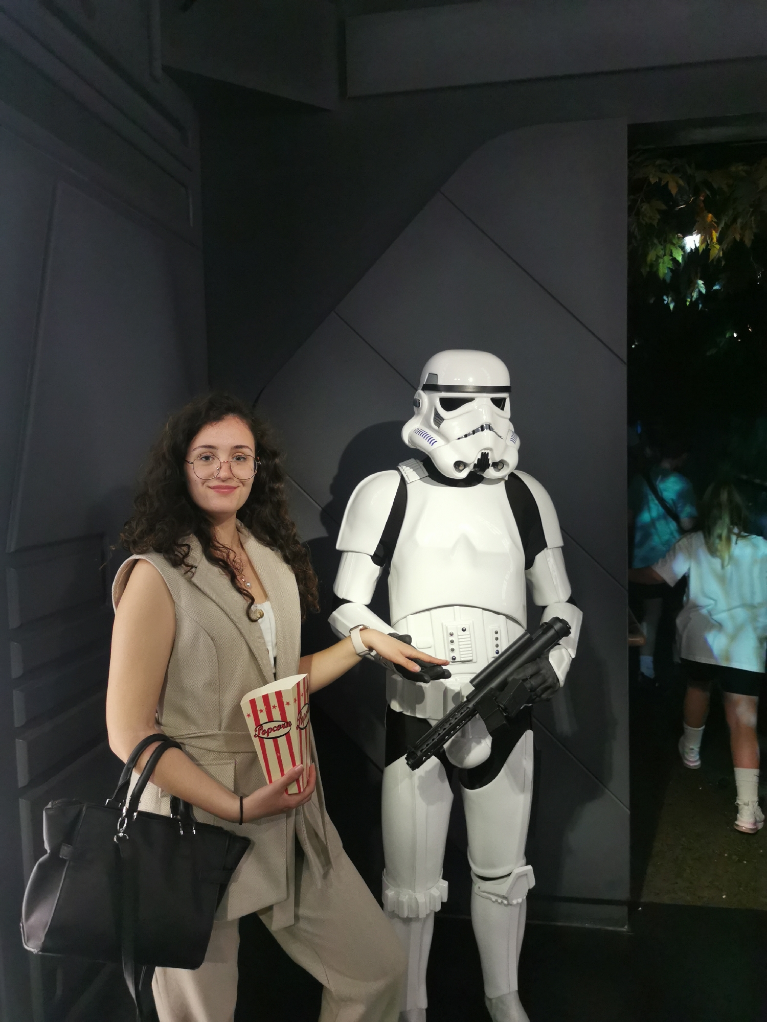 A medium shot captures a woman posing with a Stormtrooper figure. The woman, with dark curly hair and glasses, wears a beige sleeveless blazer over a white top and beige pants. She holds a black tote bag on one shoulder and a popcorn container in her other hand. She extends her arm towards the Stormtrooper.
The Stormtrooper stands rigidly next to her, holding a rifle. They are situated in a room decorated to resemble the interior of a spaceship, featuring dark gray paneling.
To the right, an open doorway reveals a glimpse of an outdoor area, where two children can be seen walking in the background. Given the Stormtrooper figure, the location is likely related to a Star Wars exhibit. The building design and vegetation visible indicate that this is in London, United Kingdom.