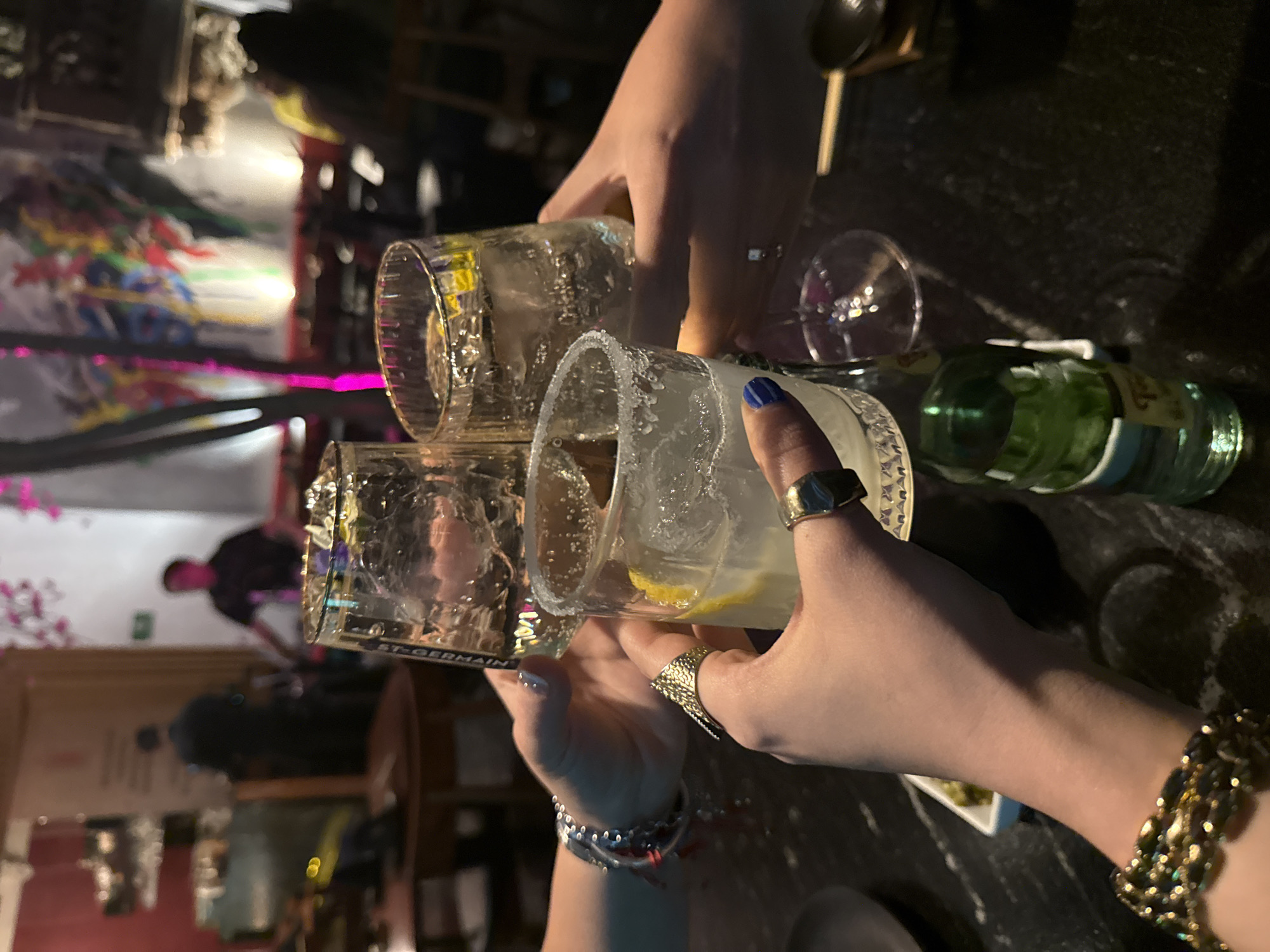 Three people are toasting with glasses of iced drinks, possibly margaritas due to the salted rim. The location appears to be a bar or restaurant, suggested by the table, glassware, and a bottle of St-Germain liqueur. There's a band performing in the background with a colorful mural on the wall. The location is likely in Mexico City, Mexico.