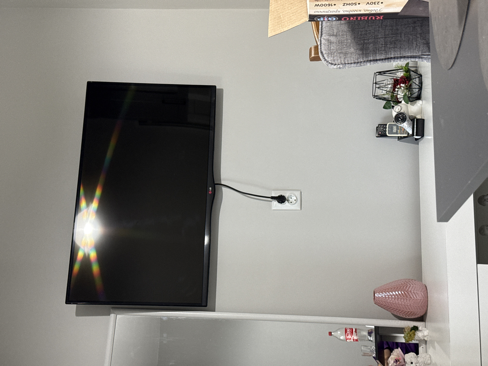 This image depicts an indoor domestic setting located in Stara Zagora, Bulgaria. A black LG flat-screen television is wall-mounted on a uniformly painted light grey wall. A power cable descends from the television to a white double electrical outlet, into which it is plugged. A significant multi-colored lens flare is present on the television screen, indicating a bright, off-camera light source reflecting from its surface.
To the immediate left of the television, a tall, white vertical structure, possibly a cabinet or part of a dresser, is visible. A mirror integrated into this structure reflects a red-capped Coca-Cola Zero Sugar bottle and a clear drinking glass. On the white horizontal surface below the mirror, a pink ceramic vase with a chevron pattern is positioned, flanked by a small white textured ornament and a miniature green and yellow floral arrangement.
On the right side of the frame, a dark grey upholstered chair with wooden armrests is partially visible. On the white surface to the left of the chair, a cluster of items is arranged: multiple remote controls (including white, black, and silver units), a small black electronic device, a white figurine wearing glasses, and a black wireframe basket containing artificial red and white flowers with green foliage. A cardboard box, labeled "RUBINO GIP" and detailing "230V • 50HZ • 1600W," is partially visible on the far right, resting on the chair.
The white furniture unit extends beneath the television, serving as a console or dresser. A dark grey tabletop surface is visible in the lower right foreground.