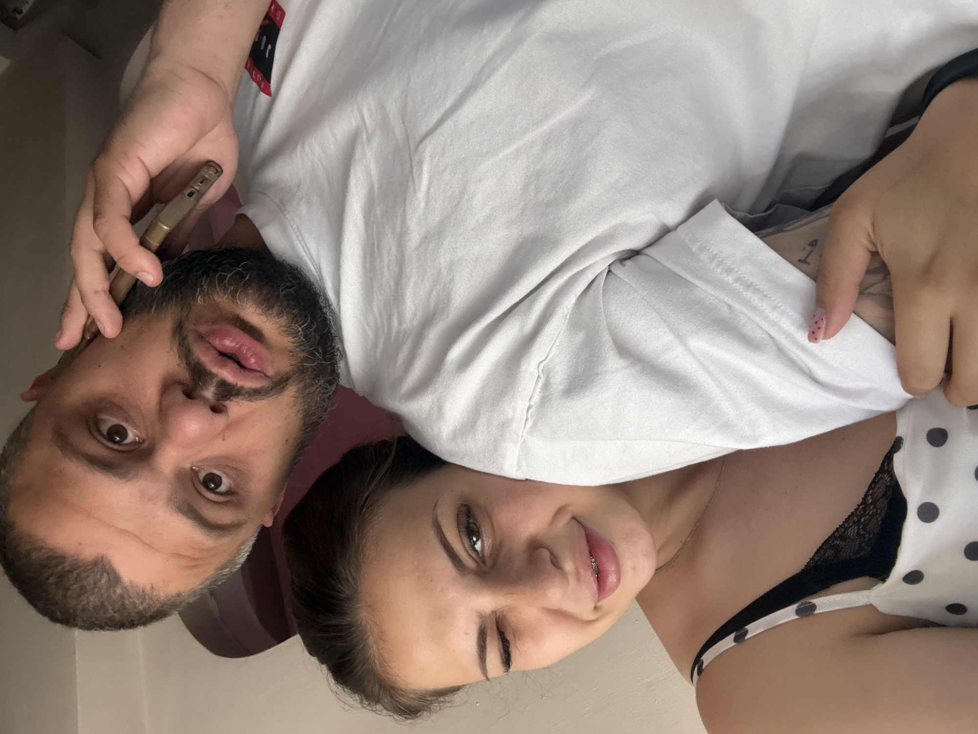 A close-up selfie shows a man on the right, holding a phone to his ear and speaking. He has a beard and is wearing a white t-shirt. To his left, a woman leans her head on his shoulder, winking at the camera. She has brown hair and is wearing a white top with black polka dots and a black lace bra. The background is indistinct, but appears to be an indoor setting, possibly a room in a home in Stamboliyski, Bulgaria. The overall impression is of a casual, intimate moment captured in a photograph.