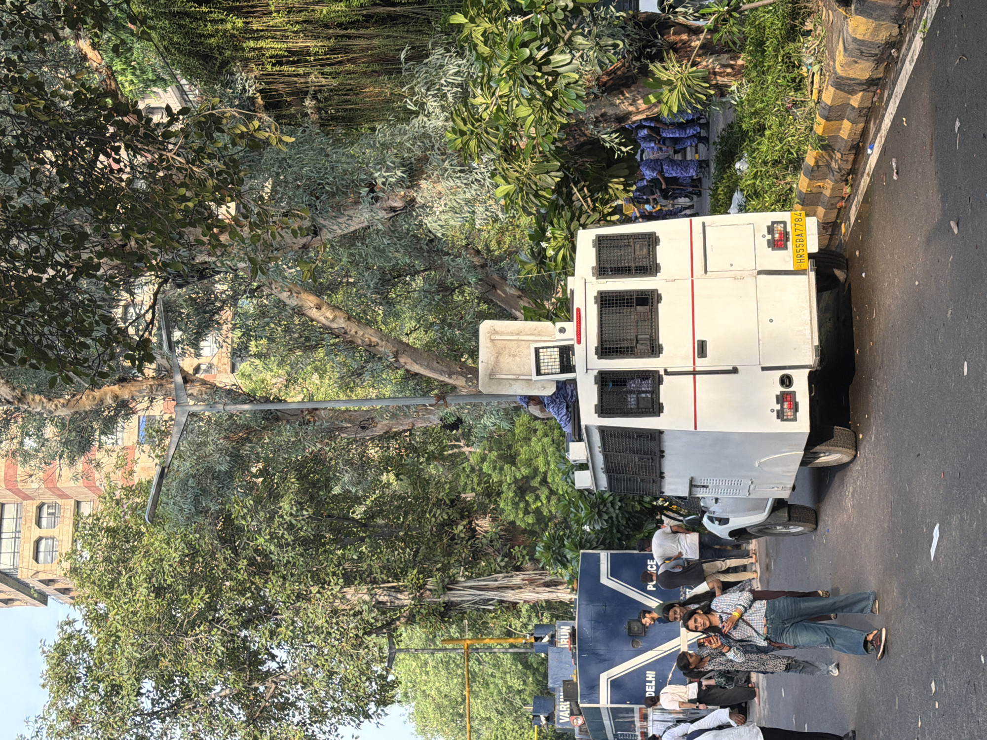 The image depicts a street scene in New Delhi, India, dominated by a large white vehicle, possibly a police or security van, with multiple caged windows. The vehicle is parked on a paved road with a yellow and black striped curb. Green trees and foliage are abundant, with one large tree in the foreground casting shadows. In the background, a multi-story building with red and beige brickwork and numerous windows is visible.
Several individuals are present. Some are standing near the white vehicle, while others are gathered around a blue vehicle with "POLICE" and "DELHI" written on its side. The people are dressed casually in various shirts, t-shirts, and jeans. Their postures and expressions do not convey strong emotions, suggesting a relatively calm or ordinary moment.
The lighting suggests it is daytime, with clear sunlight casting shadows. The presence of police vehicles and personnel implies a security presence or an event being managed by law enforcement. The overall scene appears to be an urban street, likely during daylight hours in a location that experiences warm weather, given the clothing of the individuals. The license plate on the white vehicle reads "HR55BA7784".