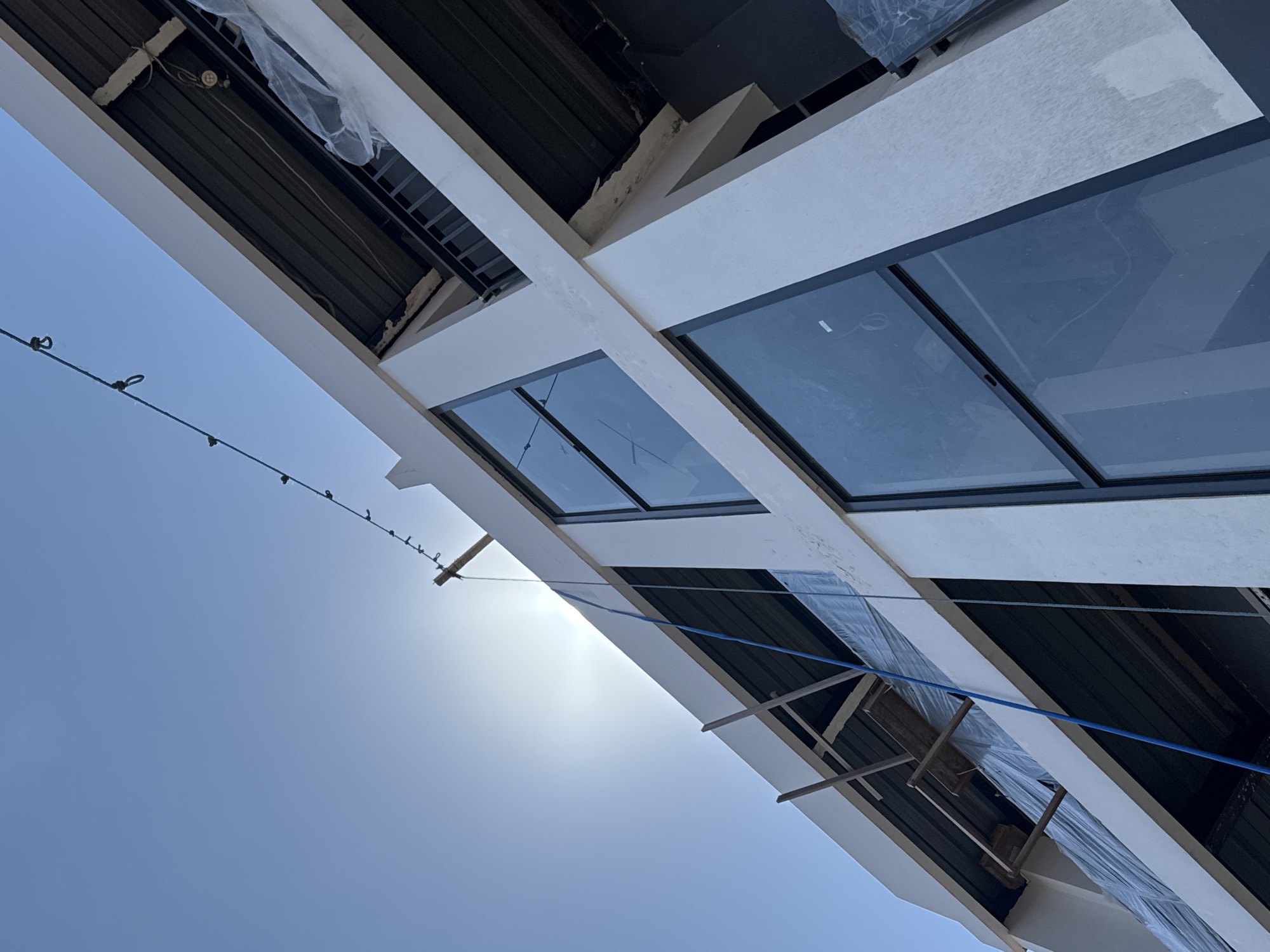 An upward-angled, wide shot of a building under construction, taken during the day. The building has large, rectangular windows reflecting the sky, and some windows are still uncovered. The building's exterior is white. Scaffolding and protective plastic sheeting are visible on the building. A string of lights is strung across the lower left corner of the image. The sky is light blue and the sun is visible through the haze. The location is Unknown, Indonesia. No people are visible in the image.