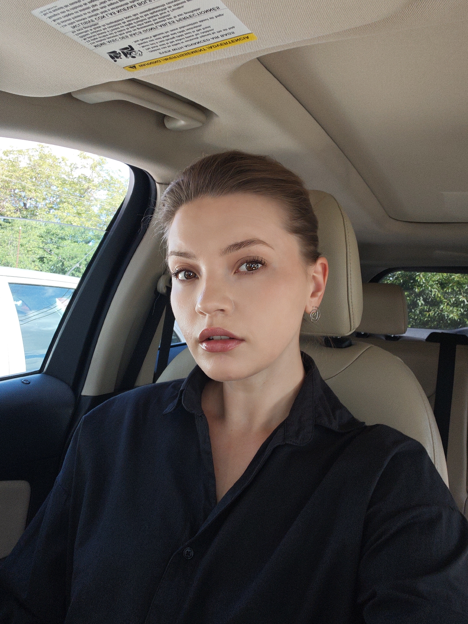 This is a close-up, indoor selfie taken from the perspective of someone sitting in the passenger seat of a car. The subject is a young woman with fair skin, brown eyes, and light brown hair pulled back from her face. She is wearing a black collared shirt. Her expression is neutral. She is wearing a small, silver, spiral-shaped earring in her left ear.
The interior of the car is visible, including a beige leather seat and a portion of the dashboard. The sun visor is also visible, and it has a label with text printed on it. Through the car windows, trees and foliage can be seen, suggesting an outdoor setting. The lighting indicates it is daytime, likely sunny.
The text on the sun visor is partially visible and appears to be warning labels or instructions.
The location context provided is Vinnytsia, Ukraine.