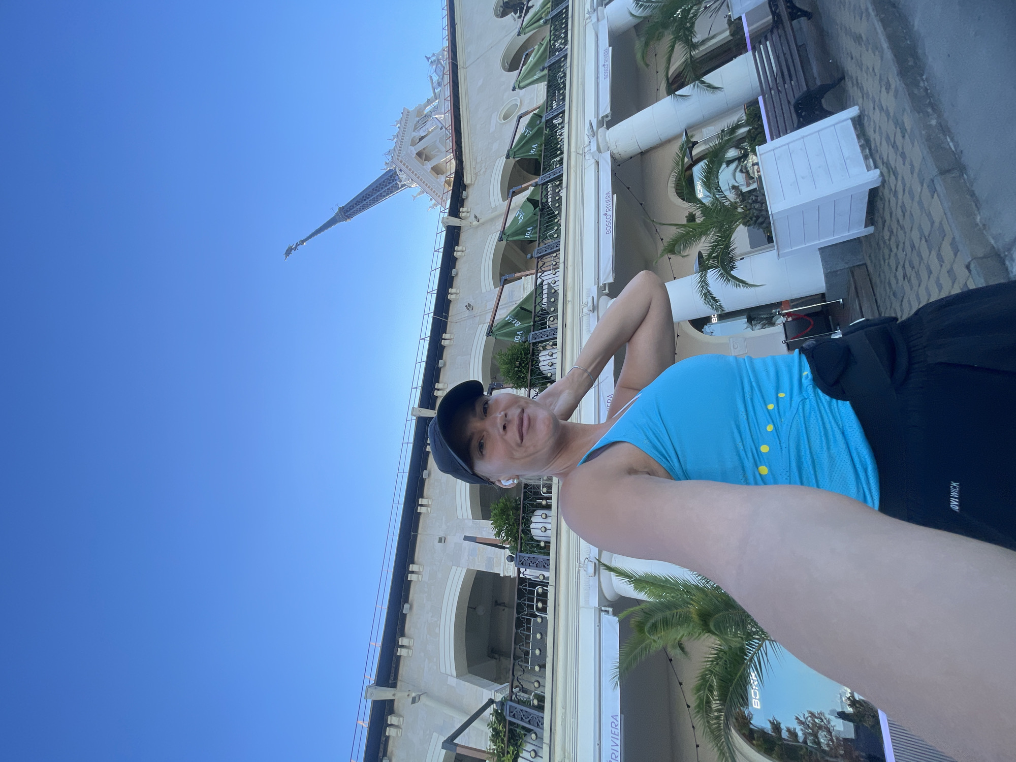 A woman in athletic wear, a blue tank top and black shorts, is taking a selfie outdoors. She is wearing a black baseball cap and white earbuds. Behind her is a large, ornate building with arches and balconies, possibly a hotel or resort. Green awnings are visible on the balconies. Palm trees are planted in white planters in front of the building. In the background, a tall, spire-like structure, reminiscent of the Eiffel Tower, pierces the clear blue sky. The ground is paved with cobblestones. The scene appears to be in a sunny, coastal location, consistent with the city of Sochi, Russia, known for its resorts and architectural landmarks.