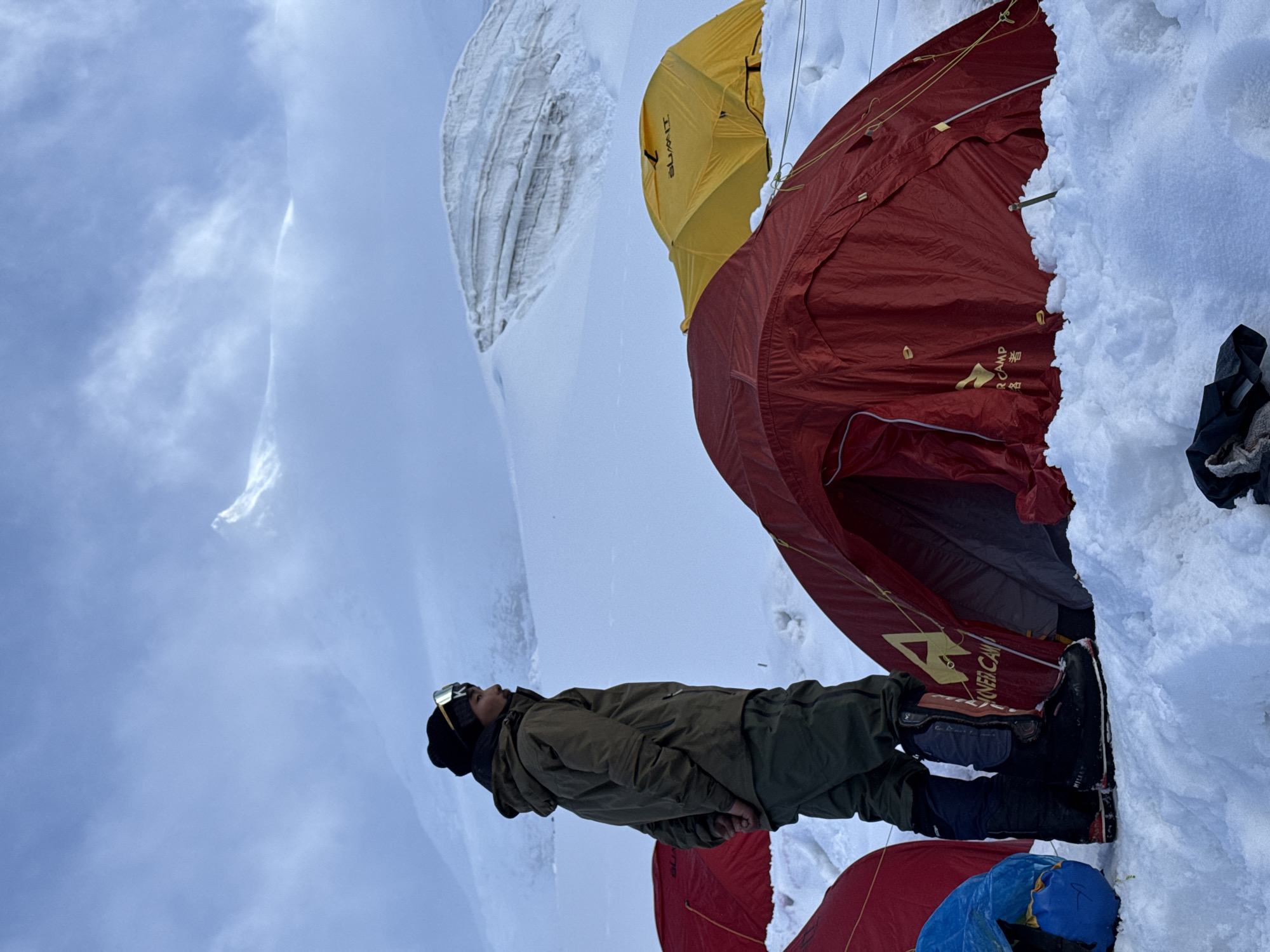 A person stands in a snowy, mountainous environment, looking upwards and to the right. They are wearing a dark beanie with goggles perched on top, a green jacket, dark pants, and heavy black boots. Behind the person, several tents are set up in the snow. Two are red, one is yellow, and a blue object, possibly a sleeping bag or pack, is visible to the left. In the background, a large, layered ice formation rises from the snow-covered ground. The sky is overcast with visible clouds, and a sharp, snow-covered peak is discernible through the haze in the far distance. The scene appears to be a campsite at a high altitude, likely associated with mountaineering expeditions in the region of Samagaun, Nepal, given the snow-covered mountains and tents.