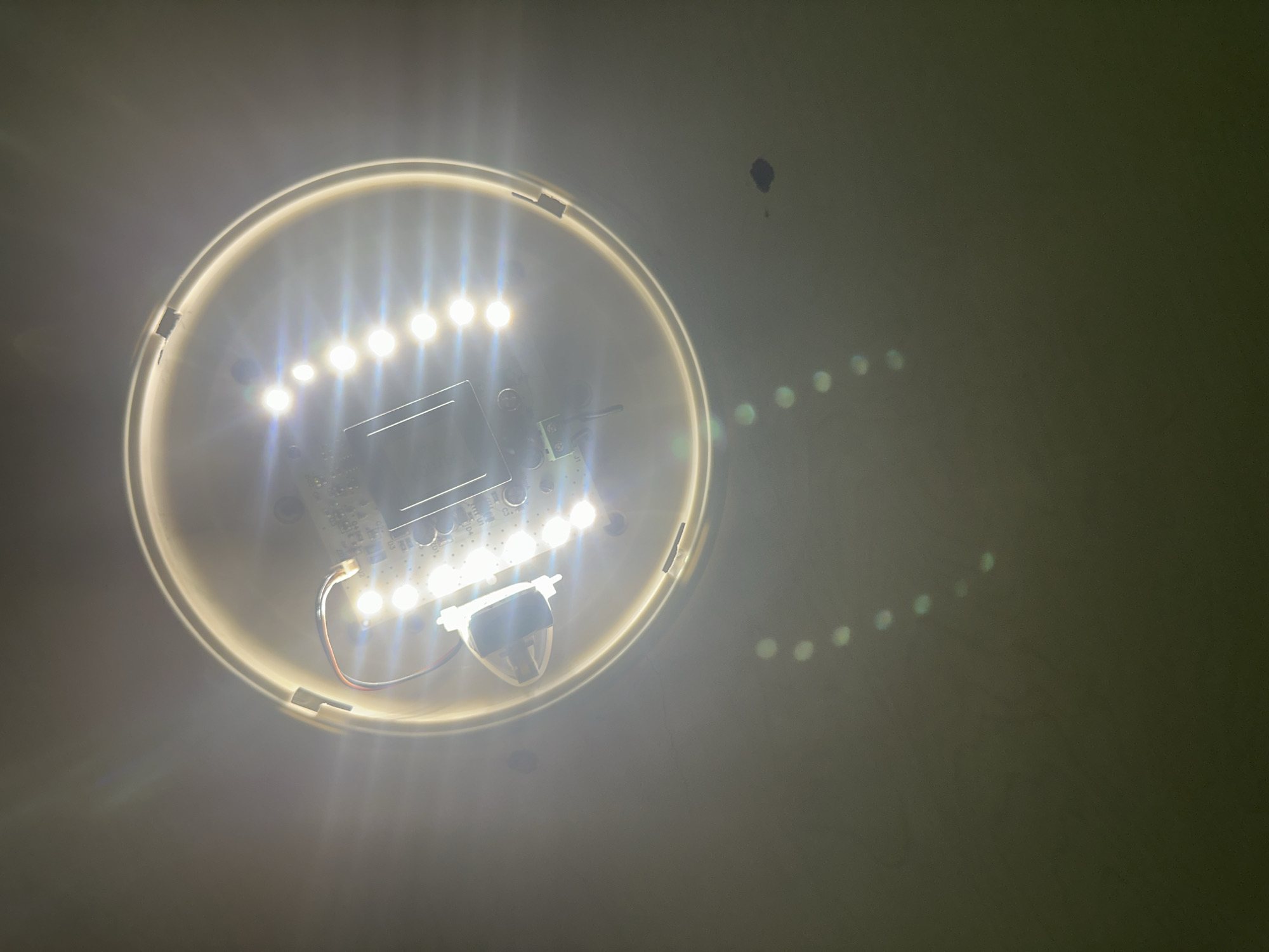 The image displays a brightly illuminated, circular LED light fixture mounted on a light-colored wall or ceiling, likely in an indoor setting in Blagoevgrad, Bulgaria. The fixture contains multiple small, round LED bulbs arranged in a ring, which are emitting intense white light. The internal circuitry of the light, including a circuit board and various components, is visible through the translucent cover. Two thin wires, one red and one blue/brown, are connected to the lower part of the fixture.
The bright light source creates prominent lens flares, appearing as a series of distinct, circular, greenish reflections that arc across the right side of the frame, moving from the center-right towards the top-right. The wall surface shows a few small, dark marks, possibly dirt or dust. The light appears to be the primary source of illumination in the scene. No people are present, and no specific activity is taking place other than the light being actively switched on. There is no visible text in the image.