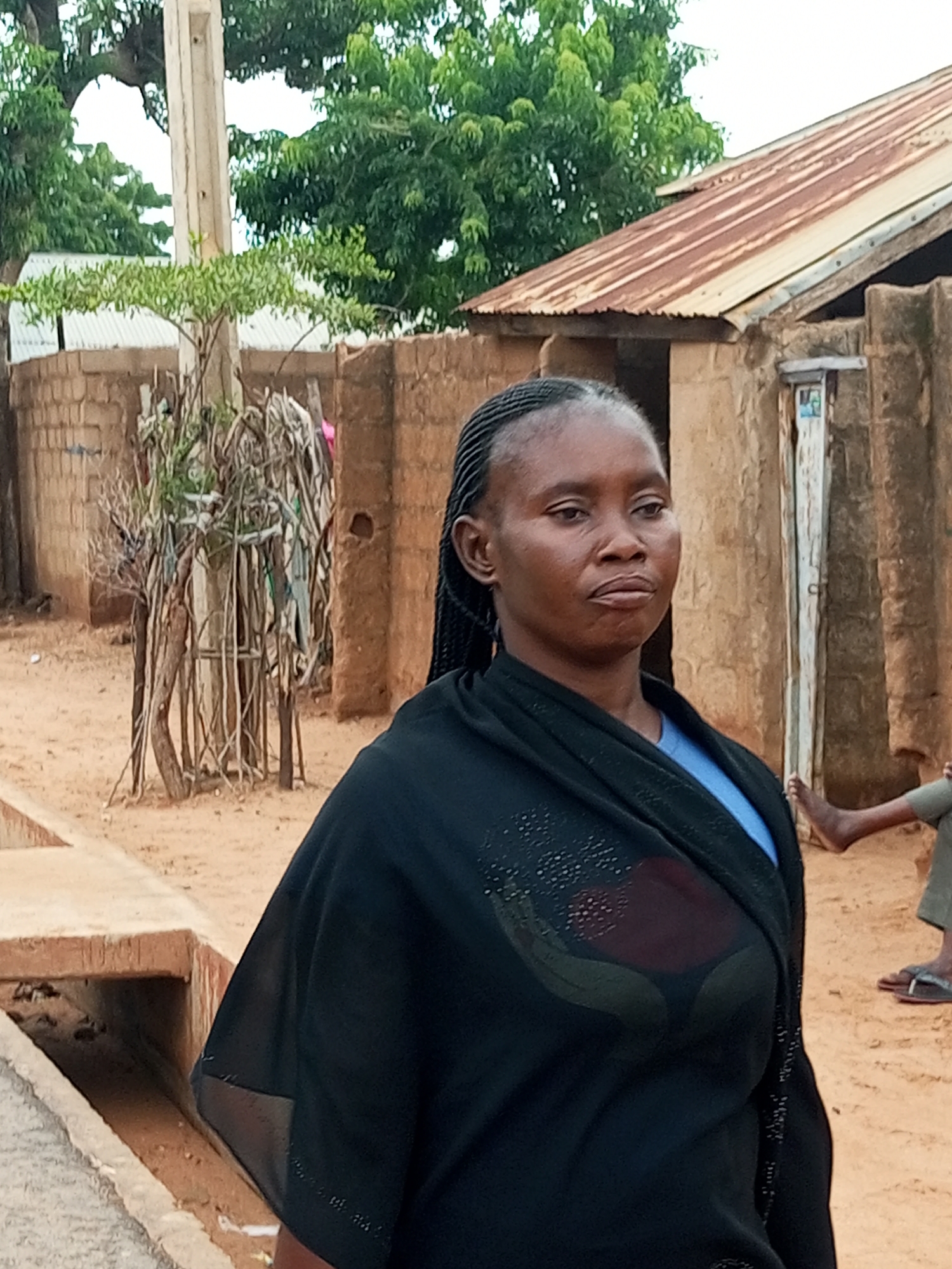 A medium shot shows a woman with dark skin and braided hair, wearing a black outer garment over a blue shirt. She is standing in what appears to be a rural or semi-urban area, with sandy ground and a concrete structure on the left. In the background, there are mud brick buildings with corrugated metal roofs, a concrete pole, and trees. To the right, another person's legs are visible, wearing sandals. The scene is outdoors, with natural light. The image quality is moderate, with some noise and pixelation, especially in the background. The overall impression is of a candid photograph taken in a simple, possibly impoverished, environment. The location is described as the city of Gombe, Nigeria.