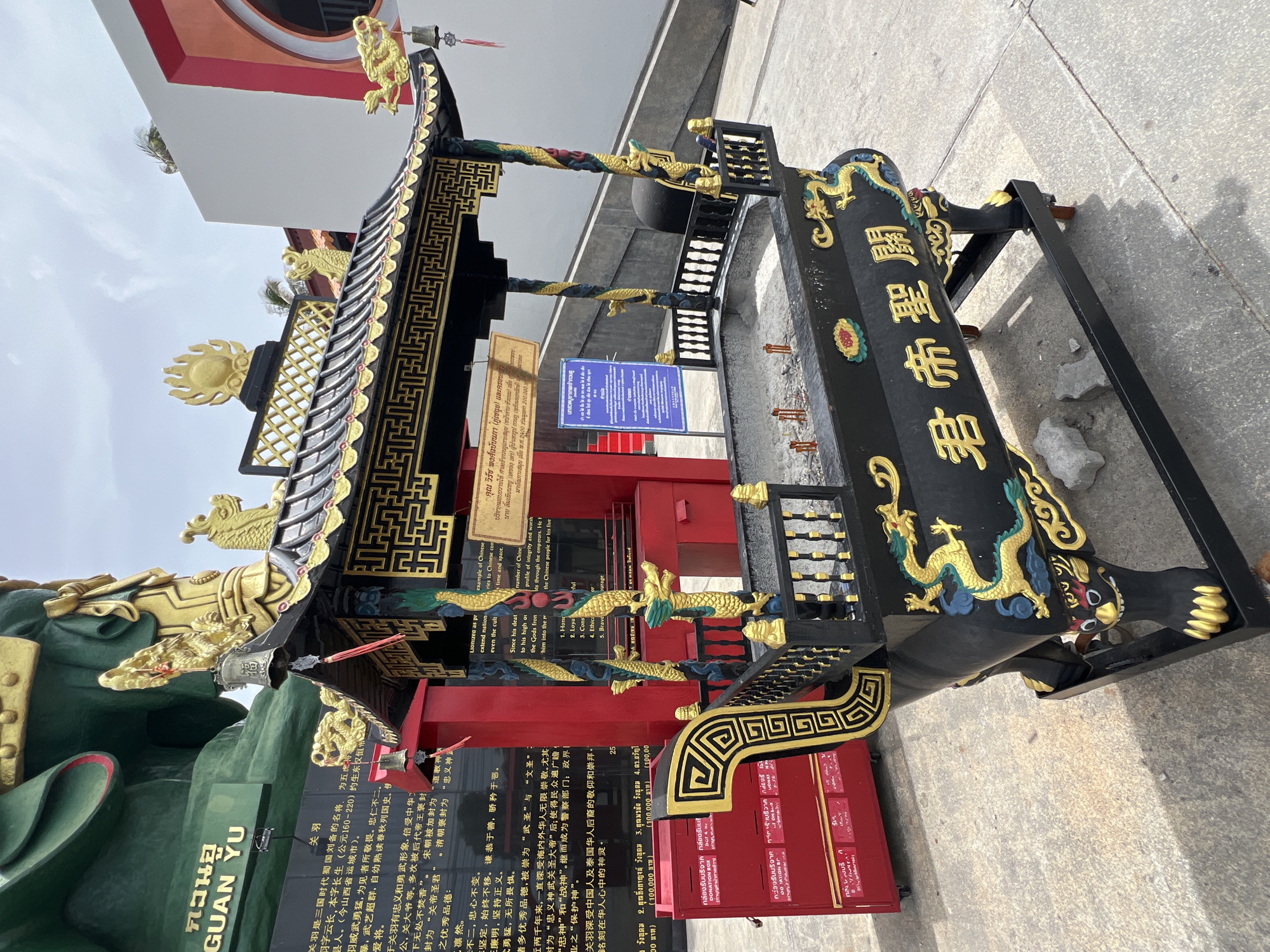 In Baan Hua Thanon, Thailand, an ornate black incense burner with gold dragon motifs sits on a concrete surface in front of a large green statue. The burner features a tiered, black and gold roof adorned with smaller dragons and decorative elements, and a railing around its top. Inside the burner, remnants of burnt incense sticks are visible. To the left of the burner, a bright red donation box is placed on the ground, with Chinese and Thai text displayed on its surface. Behind the burner and donation box, a dark red structure with stacked red boxes and a visible staircase can be seen. Further in the background, a section of the large green statue with the name "GUAN YU" inscribed in white text on a dark green plaque is visible. The sky above is overcast with scattered clouds.