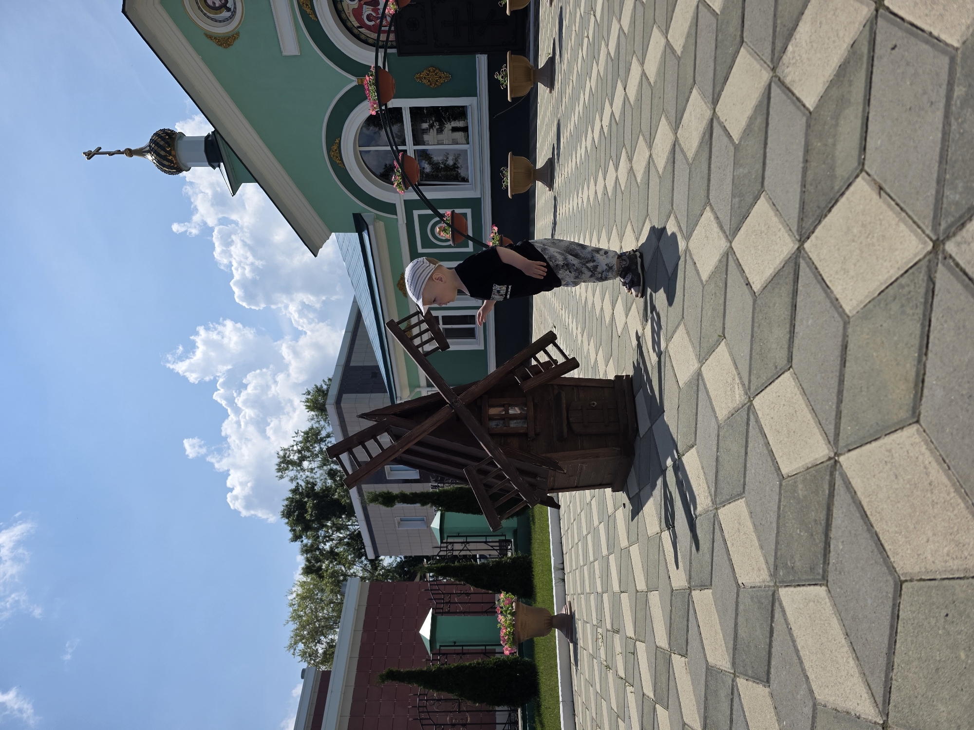 The image depicts a young child, wearing a black t-shirt, camouflage pants, and a striped cap, standing on a patterned paved area. The child is facing a small, wooden windmill model and appears to be interacting with it. In the background, a green building with a gold-domed roof and a cross is visible, suggesting it is a church or religious structure. Several potted plants are placed around the building and on the pavement. The sky is blue with scattered white clouds. The environment appears to be an outdoor public space, possibly a park or courtyard within the city of Chaikovsky, Russia.