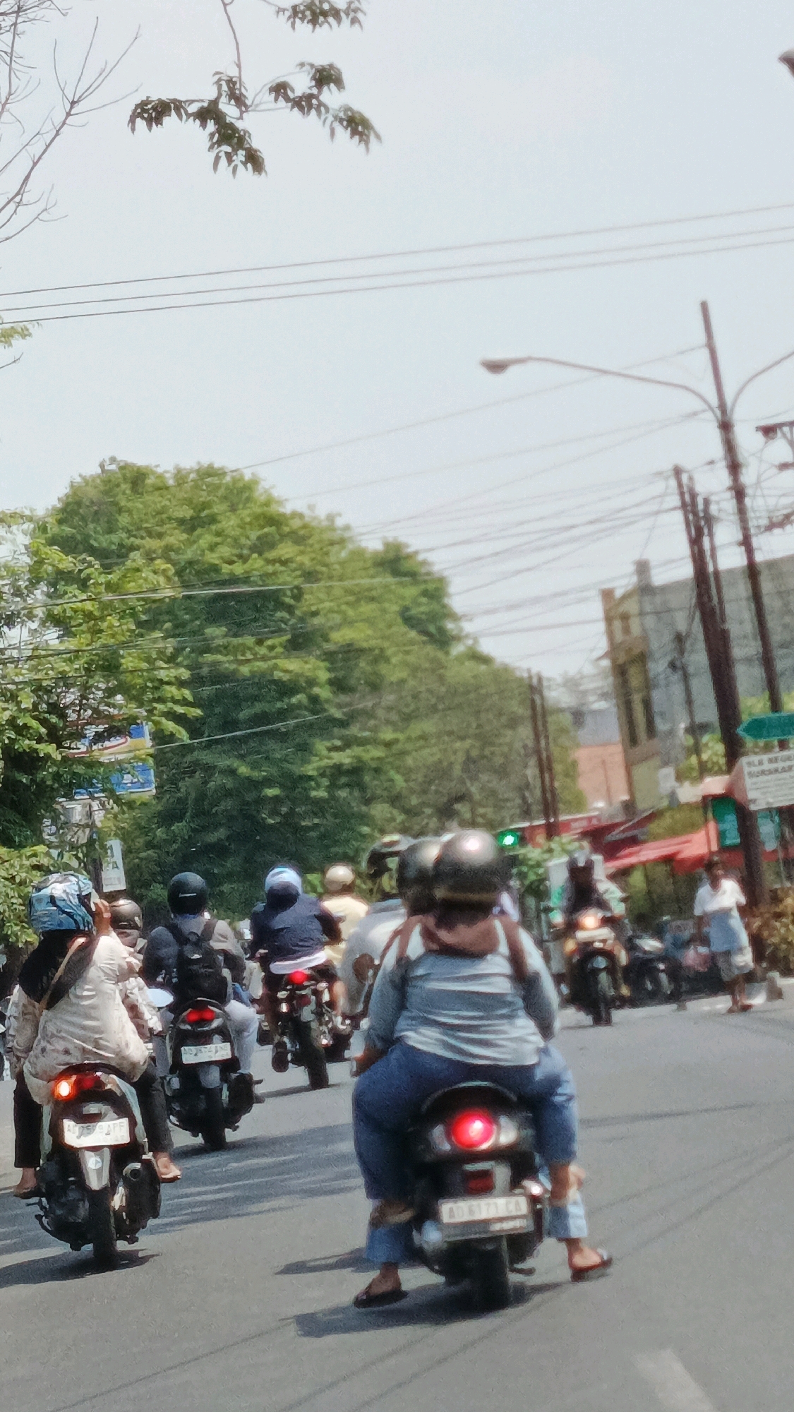 The image depicts a street scene in the city of Surakarta, Indonesia, with multiple motorcycles and riders navigating a road. The foreground shows the rear of two motorcycles, one on the left with a rider wearing a patterned headscarf and a light-colored top, and another on the right with a rider in a blue top and jeans. Both motorcycles have illuminated red taillights. The road surface appears to be asphalt.
In the mid-ground, several other motorcycles and riders are visible, some with helmets and varying attire. Trees with lush green foliage line the left side of the road, and in the background, buildings and overhead power lines are present. A traffic light, showing green, is visible on the right side of the road. The sky is overcast and bright. The overall impression is a busy urban street with typical daily traffic.