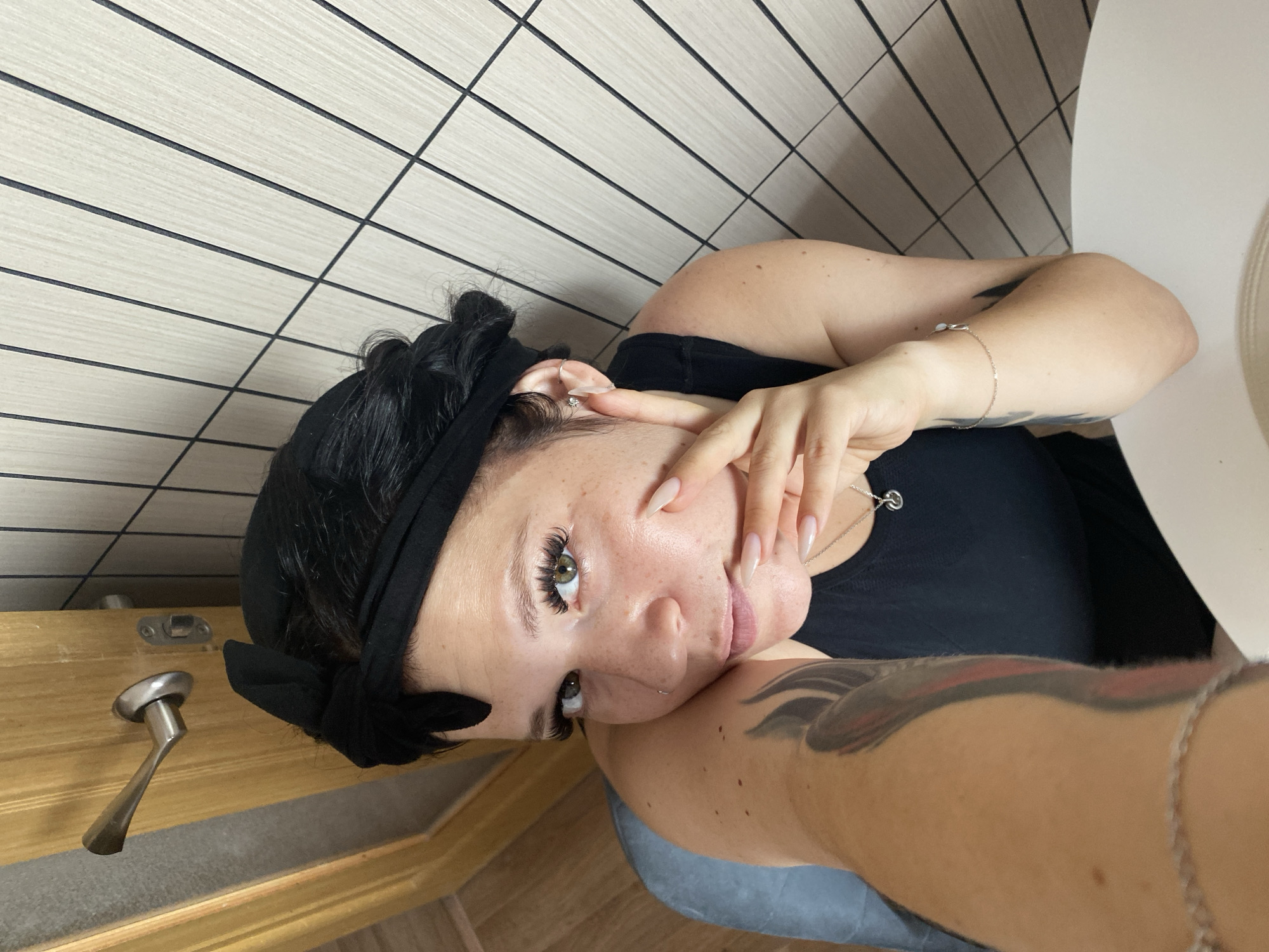 A woman with dark hair tied back with a black fabric headband is shown from a high angle, looking at the camera. She has on a black tank top and is wearing earrings and a necklace with a small pendant. Her left arm is extended towards the camera, revealing a tattoo. Her right hand is resting on her chin. The background consists of a wooden door with a metal handle on the left and a tiled wall with vertical dark lines on the right. The lighting is bright, suggesting an indoor setting.