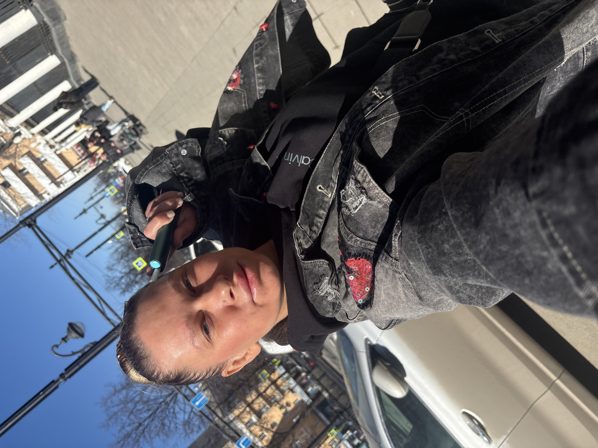 A woman is taking a selfie outdoors. She is holding a black, cylindrical object to her face. She has light skin and her hair is pulled back. She is wearing a black hoodie with a white collar under a dark gray denim jacket. The jacket has red patches. A silver car door is visible to her right. The background shows a street scene, with buildings, trees, and street signs, which point to the city of Perm, Russia, as the location.