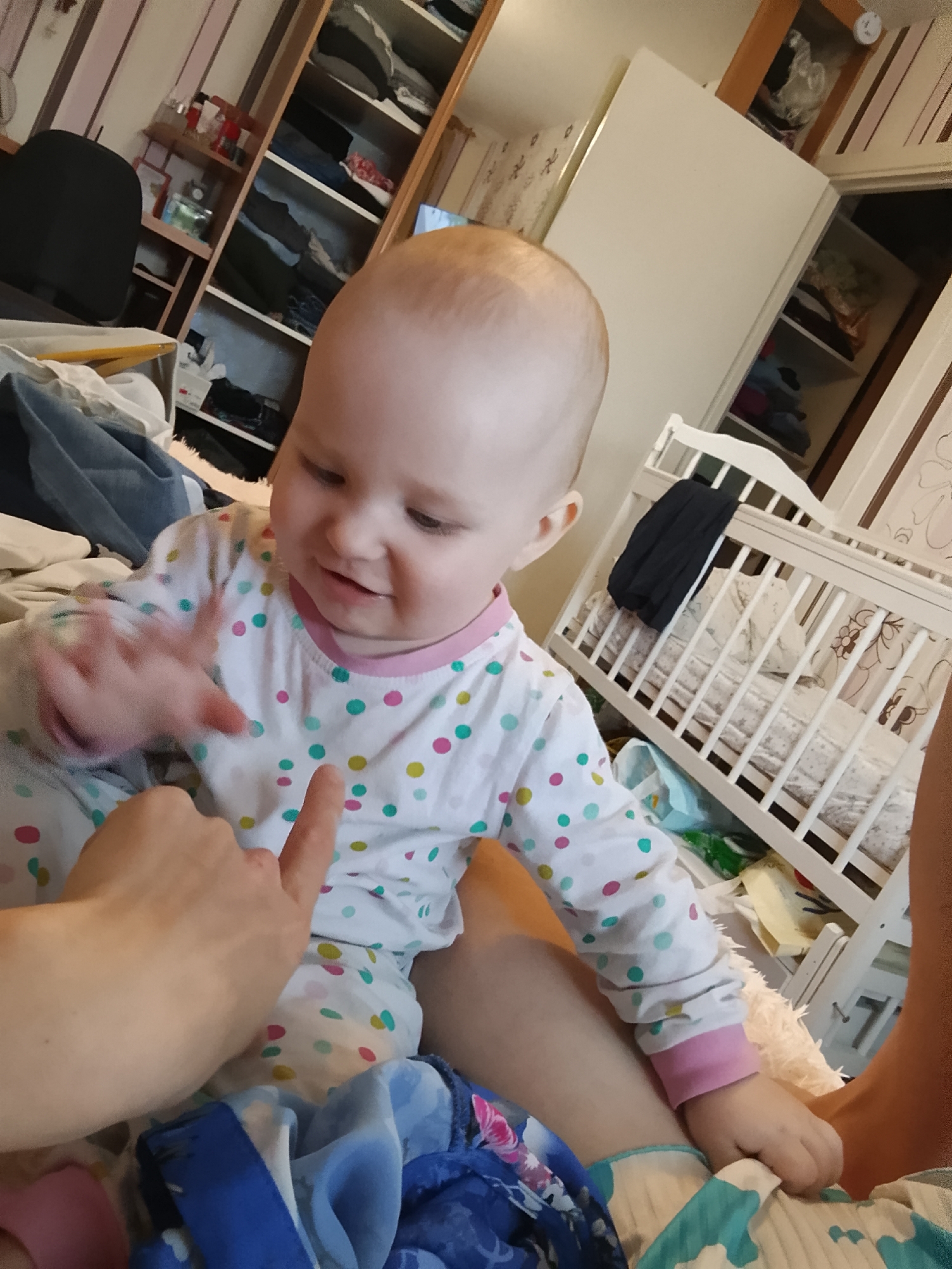A child is being held by an adult in what appears to be a bedroom or play area. The child, wearing pajamas with polka dots, is looking at the adult's finger, which is extended towards the child. The adult's hand is also visible. In the background, there is a crib with bedding and a black garment draped over it. Shelving units filled with clothes and other items are visible on the left side of the image. The overall setting suggests a domestic environment. The image was taken in Minsk, Belarus.