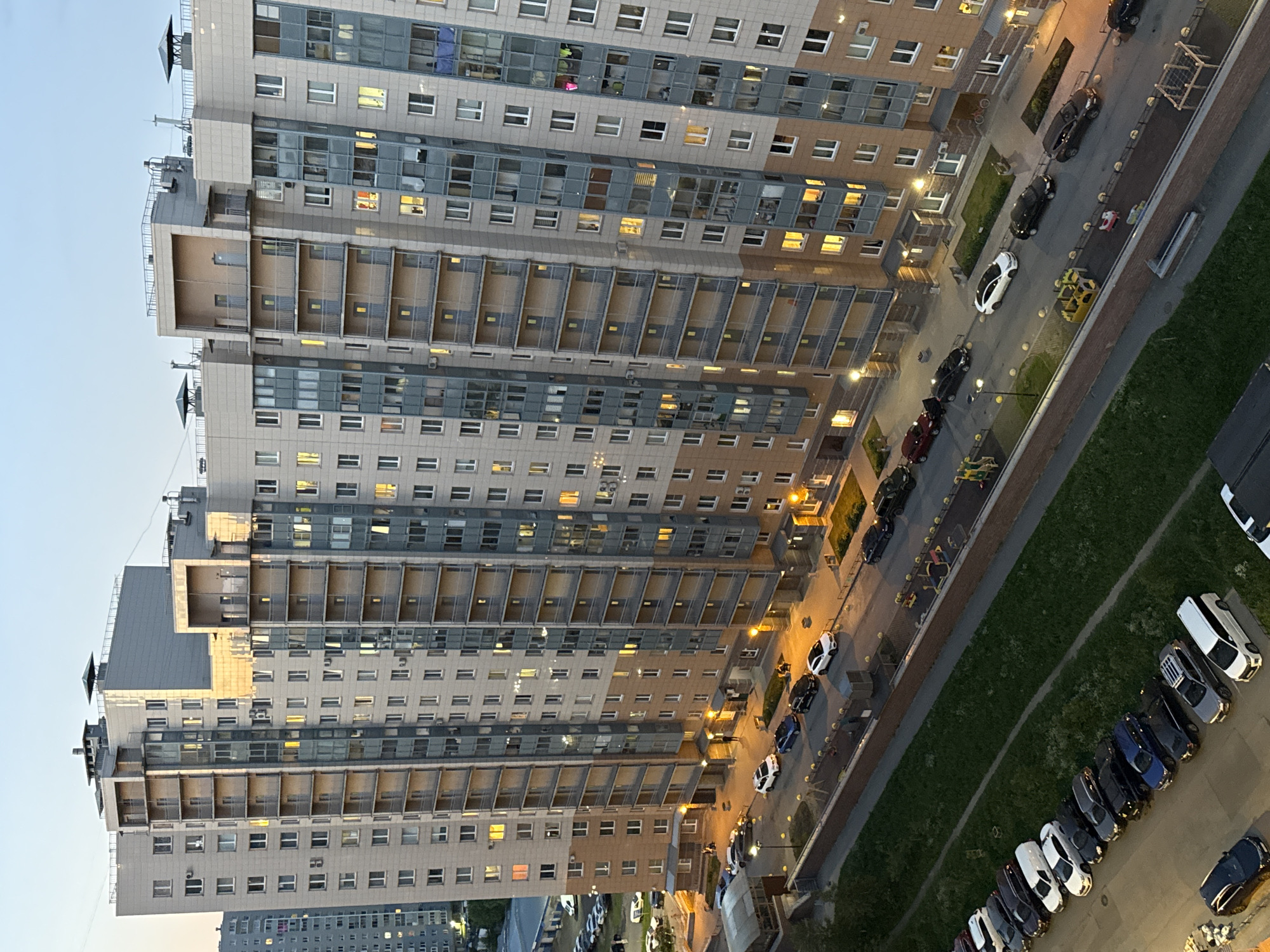 The image depicts a high-angle view of a contemporary residential district in an unknown city in Russia at dusk. A prominent, elongated multi-story apartment building with a light grey upper facade and a brown lower section dominates the scene. The building features numerous rectangular windows, many illuminated from within, and multiple recessed balconies with metal railings or enclosed glass panels. A taller, older grey apartment building is partially visible in the background to the left.
At street level, a paved road runs parallel to the main building, lined with parked automobiles including sedans, SUVs, and a white minivan. Several individuals are observed on sidewalks: one person walks away from the camera near the center, and two people are seated on a bench to the right. Streetlights cast yellow illumination on the road and pedestrian areas. Multiple children's playgrounds, equipped with swings, slides, and climbing structures, are situated along the street and between buildings. Green spaces with grass and a few trees are visible in the foreground and between structures. Bollards separate the roadway from pedestrian zones and playgrounds. A larger parking lot containing additional vehicles is visible to the far left. The overall scene indicates a populated, organized urban residential environment.