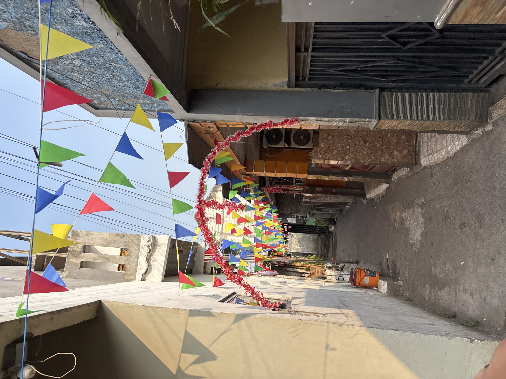 The image depicts a narrow alleyway decorated with colorful triangular flags strung across it, creating a festive atmosphere. Red tinsel or garland is also draped overhead, forming an archway. Buildings line both sides of the alley, with one side appearing to be a residential or commercial structure with visible air conditioning units. The other side shows a building with textured walls and a window. At the entrance of the alley, a bright orange bucket sits on a concrete block. The ground is paved with asphalt. In the background, the alley continues into the distance, with more buildings and decorations visible. The sky above is clear and blue, suggesting daytime. The scene is likely in a densely populated urban area, and the decorations suggest a celebration or special event. The specific location in Bekasi, Indonesia, is characterized by this type of urban setting with narrow passageways and closely situated buildings.