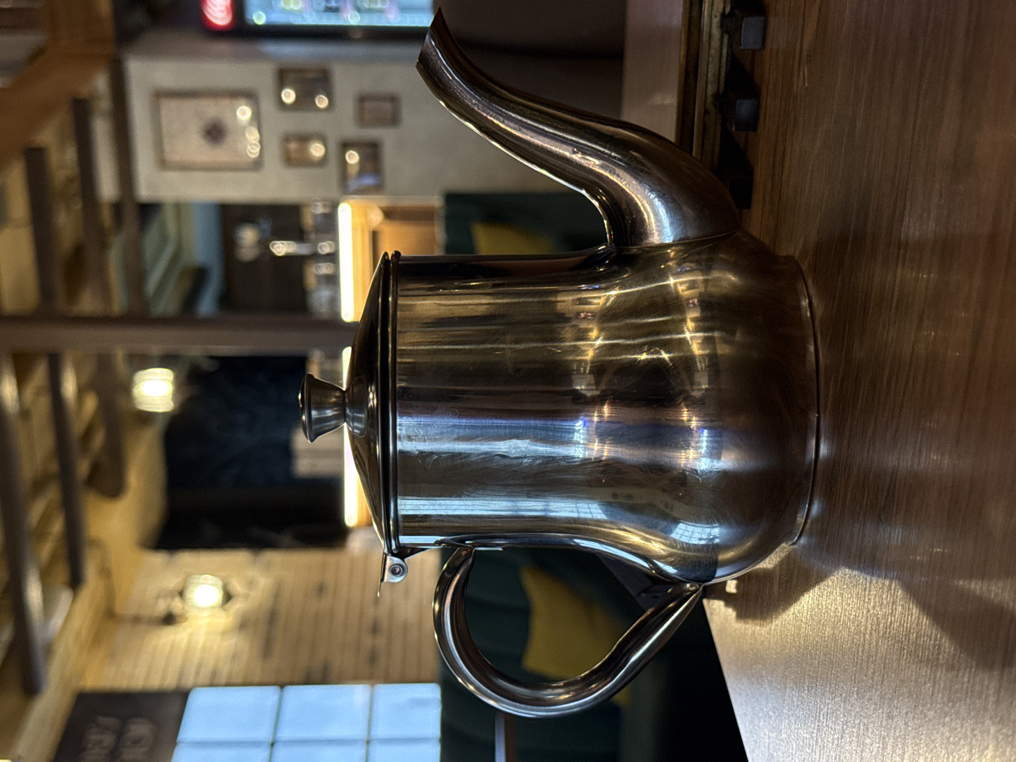 A polished metal teapot is centered on a wooden table, reflecting the warm interior of a cafe. The background shows the cafe's ambiance with exposed wooden beams on the ceiling, light fixtures, and framed pictures on the wall. There are also glimpses of seating and a Coca-Cola display. The scene suggests a quiet moment within this establishment. This image was captured in Krasnodar, Russia.