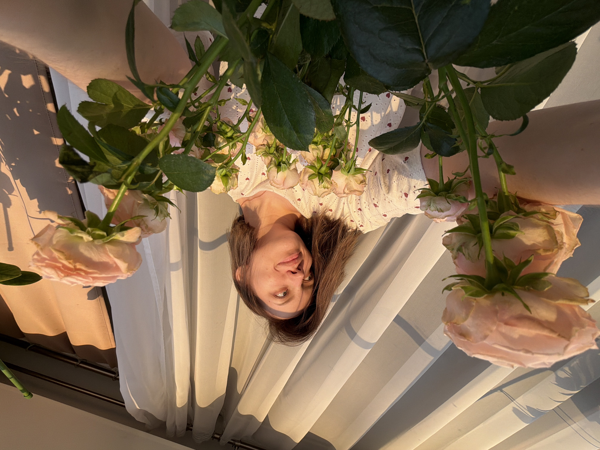 The image is a creatively composed portrait of a young woman lying down, looking upwards at the camera, with a bouquet of light pink roses positioned above her face and upper body. Her head, with dark brown hair, is centrally located in the frame. She exhibits a calm, open expression with a subtle, gentle smile. She is wearing a light-colored garment, possibly a top or dress, adorned with small heart-shaped cutouts or embroidery.
The roses feature delicate, pale pink petals and prominent green stems and leaves, some of which show signs of slight fading or dryness, contributing to a natural, organic aesthetic. Multiple rose blooms and buds are visible, creating a soft framing around her face.
The background features flowing curtains. To the viewer's left, there is a section of a thicker, beige or light brown curtain, which casts elongated, warm shadows onto a lighter, possibly sheer white curtain on the right. The soft, warm, golden light and the distinct shadows strongly suggest the photograph was taken during the golden hour, likely late afternoon. The overall atmosphere is gentle, serene, and natural. No text is visible in the image. The setting appears to be an indoor space, possibly a home in Reshetnikovo, Russia, though no explicit location cues are present.