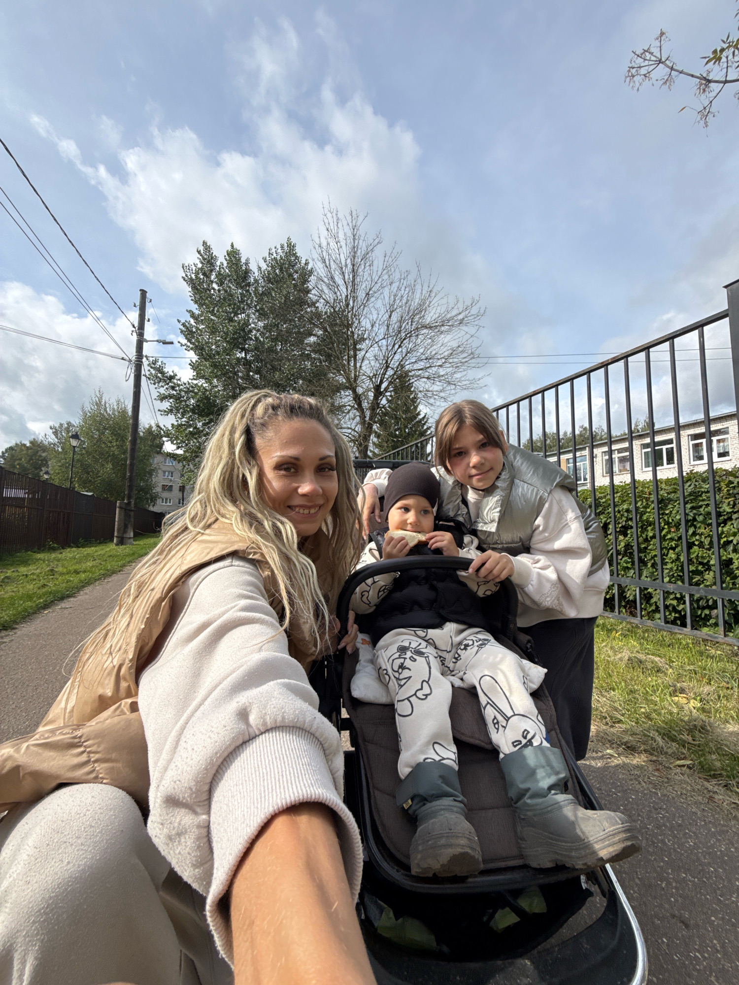 In a selfie taken outdoors, a blonde woman with braided hair smiles at the camera, holding a stroller. Next to her, a young girl in a puffy vest stands and looks at the camera. Inside the stroller, a toddler wearing a beanie and patterned pants is eating a piece of bread. The background shows a fence, a grassy area, trees, a power pole with wires, and buildings in the distance. The scene appears to be in a residential area.