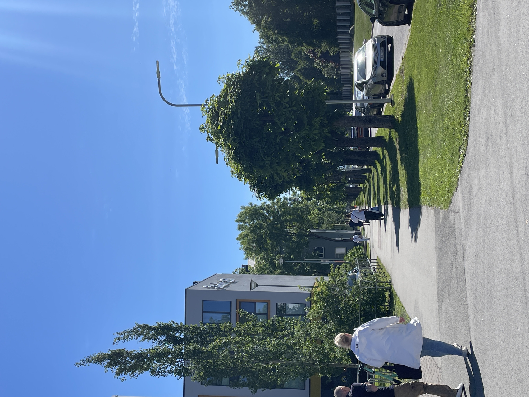 The image depicts a sunny day in Tallinn, Estonia, with a clear blue sky. A paved pathway runs alongside a grassy area lined with trees. Several cars are parked on the right side of the pathway, and a modern building with large windows is visible on the left.
There are several people in the image, some walking along the pathway. In the foreground, a person wearing a white lab coat and blue jeans is walking away from the camera. Further down the path, a group of people, some in white attire, are also walking. The presence of people in lab coats suggests a possible connection to a medical or research facility.
The scene appears to be an outdoor area of a campus or a public space. The sunlight indicates it is daytime, likely late morning or afternoon. The lush green trees and grass suggest a temperate season, possibly spring or summer. The architecture of the building and the style of the cars do not provide any particularly strong clues to a specific era, but the overall impression is modern.
No distinct text is visible in the image, apart from what might be branding on the clothing of the people, which is too indistinct to read. The overall mood is peaceful and ordinary, with people going about their day under clear skies.