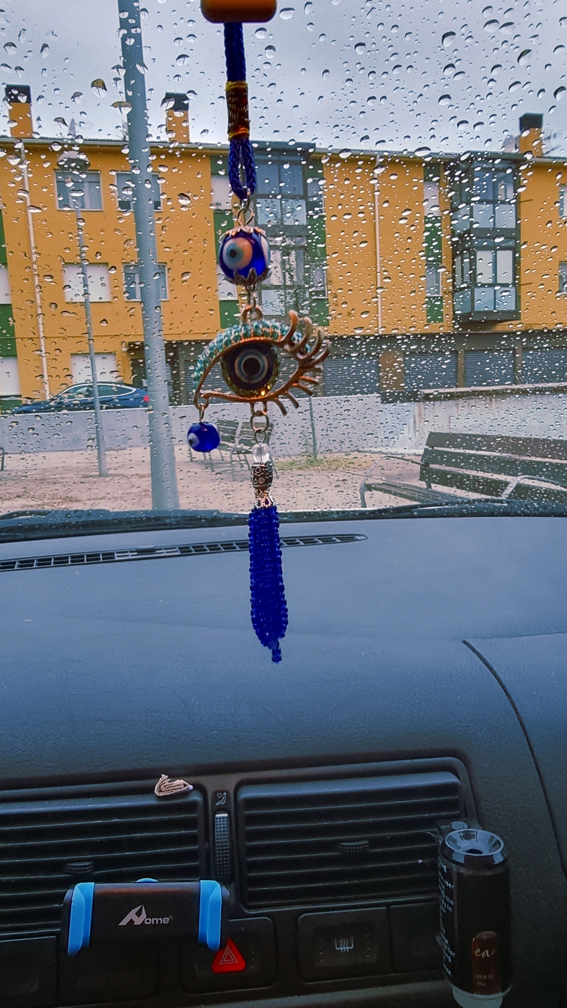 The image shows a close-up view of a car's interior dashboard on a rainy day. A blue beaded evil eye ornament is hanging from the rearview mirror, obstructing the view of a yellow and green building with multiple windows and balconies, which is part of the city of Sant Joan Les Fonts, Spain. The windshield is covered in raindrops. The dashboard features air vents, a hazard light button, and a car phone holder. A dark-colored air freshener can is also visible. The lighting is dim, suggesting an overcast sky.