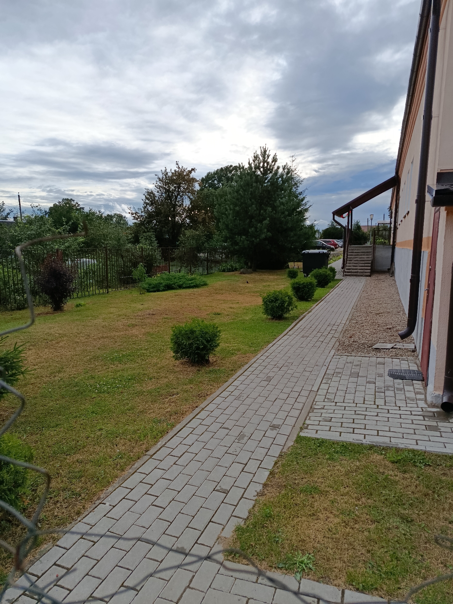 The image captures a view of a paved walkway leading alongside a building with light-colored walls and a dark-brown downspout. The walkway is constructed of gray rectangular pavers. To the left of the walkway is a grassy area with scattered bushes and a metal fence bordering it. Beyond the fence, there are trees and foliage. The sky is overcast with clouds. In the distance, vehicles are parked near the building. The scene appears to be an outdoor area adjacent to a residential or commercial property. The location is identified as Смілавіцкі сельскі Савет, Belarus. There are no people visible in the image.