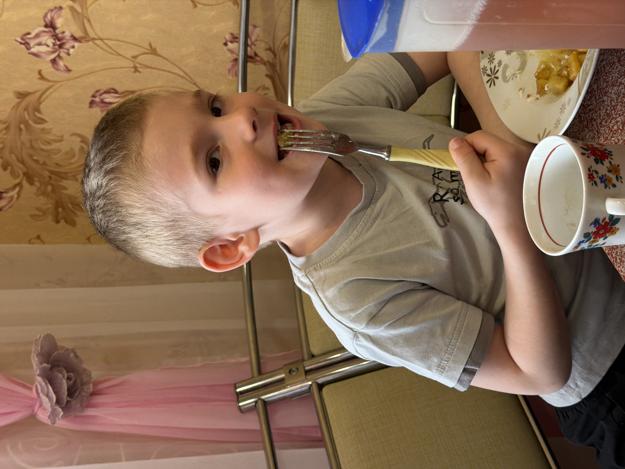A young boy with short, light-colored hair is captured mid-meal in an indoor setting. He is wearing a light-colored, short-sleeved t-shirt, which has an unidentifiable dark graphic or text on the left chest area. The boy is actively eating, holding a fork with a light-colored handle to his mouth, which contains some diced, light-colored food, possibly stewed fruit or vegetables. He is looking directly towards the camera with a neutral expression.
On the table in front of him, there is a white plate containing more of the diced food. Next to the plate is a white mug adorned with a colorful floral pattern and a red rim. A clear plastic container with a blue lid and reddish liquid, likely a drink, is also visible. The boy's right arm is bent at the elbow, resting near the mug.
The background features patterned wallpaper with a repeating motif of brown/gold foliage and purple flowers. A pink fabric, possibly a curtain or tablecloth, is seen in the lower left, decorated with a grey/purple fabric flower. Parts of a metallic furniture frame, likely a chair or table, are also visible. The scene suggests a casual mealtime at home in Засечное, Russia.