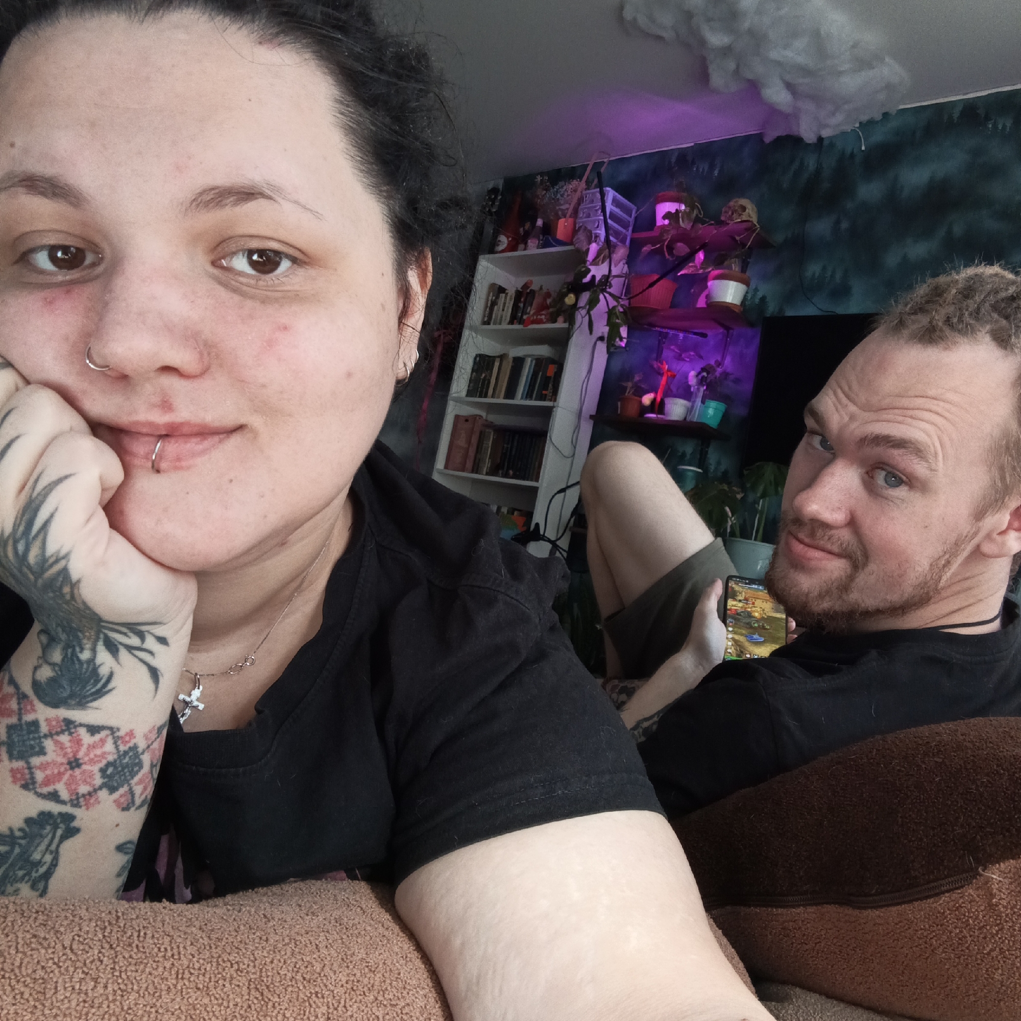 This image depicts two individuals, a female and a male, in a casual indoor setting within the city of Chaikovsky, Russia.
The female is prominently positioned in the foreground, facing the camera with a slight smile. She has dark hair, a fair complexion with some minor blemishes, brown eyes, a silver nose ring, and a silver lip ring. Her right hand, resting under her chin, exhibits extensive dark-ink tattoos covering the back of her hand and forearm, including patterns resembling a rooster and a red-and-black geometric design similar to cross-stitch. She wears a black t-shirt and a silver chain necklace with an infinity symbol and a crucifix pendant. She is seated on a light brown, textured fabric surface.
Behind and to the right of the female is a male, also looking towards the camera with a slight smile. He has light brown, possibly dreadlocked or matted hair, a beard, light-colored eyes, and visible wrinkles on his forehead. He wears a black t-shirt and dark shorts, with one leg visible. He is holding a smartphone in his right hand, displaying a colorful video game interface. He is resting on a darker brown, textured fabric surface.
The background features a wall covered in dark teal wallpaper with a black forest silhouette pattern. A white multi-shelf bookcase filled with books is visible on the left. To the right, several floating shelves hold potted plants, some illuminated by a distinct purple light, alongside other decorative items including a white skull-like object. A large, white, cloud-shaped decoration is affixed to the light-colored ceiling. Electrical cords are visible running along the wall and shelves.