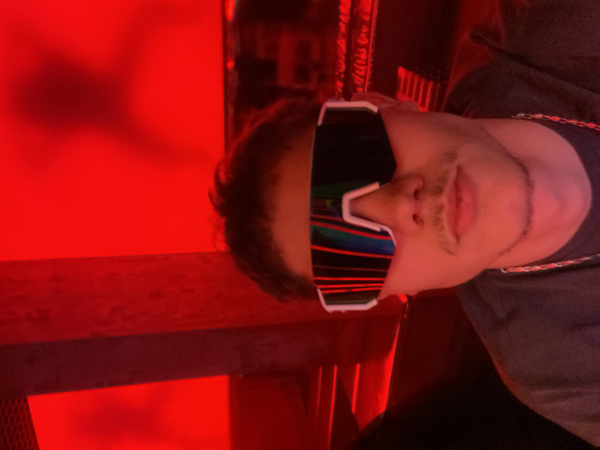 A man wearing large, futuristic white sunglasses with a reflective, striped lens is positioned in the foreground of a vertically oriented photograph. The image is dominated by red lighting. The man has short brown hair and a thin mustache. He is wearing a gray shirt and a silver chain around his neck. The background includes blurred elements of metal and what appears to be a grated surface, all rendered in deep shades of red. The image's orientation suggests it was taken by a mobile device and may have been rotated. There is no information available to place the subject at any location in the city of Sofia, Bulgaria.